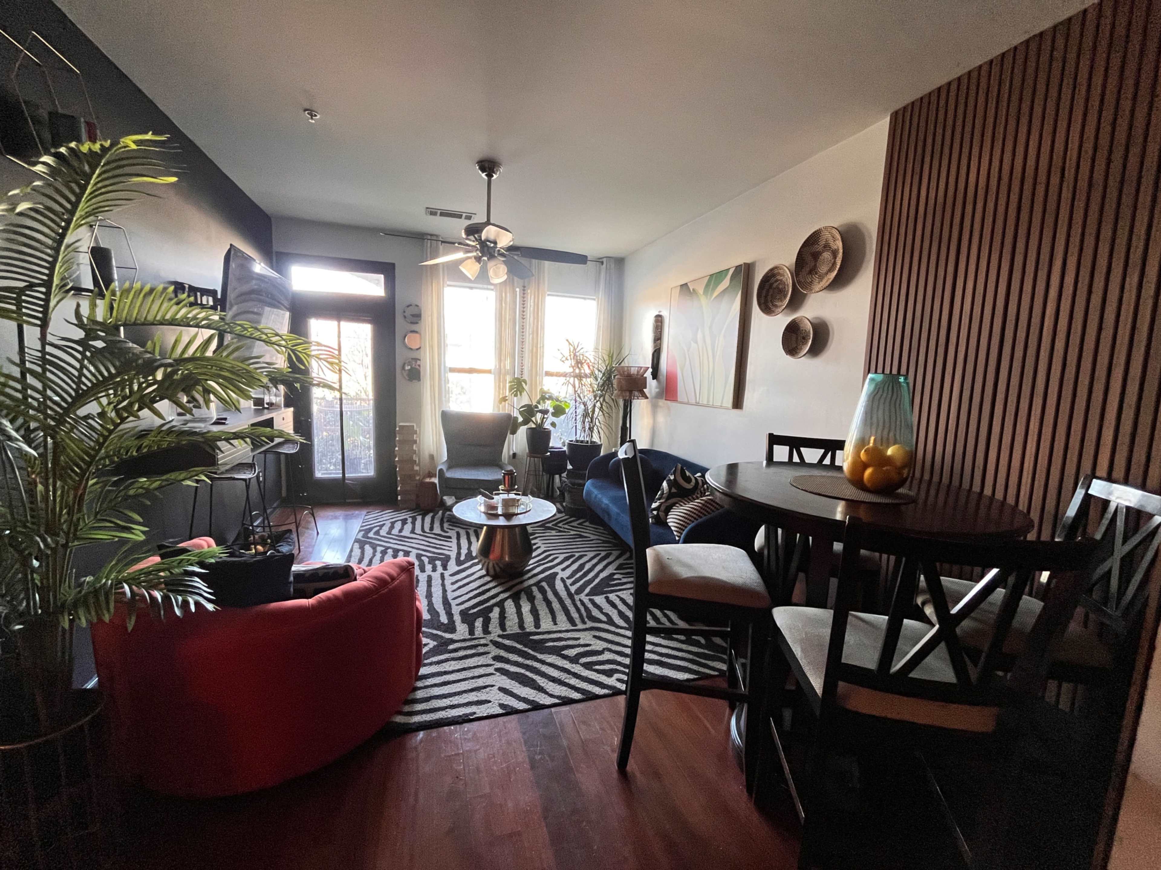 The image shows a stylish living area with a mix of seating options, decorative plants, a dining table, and a large window allowing natural light.