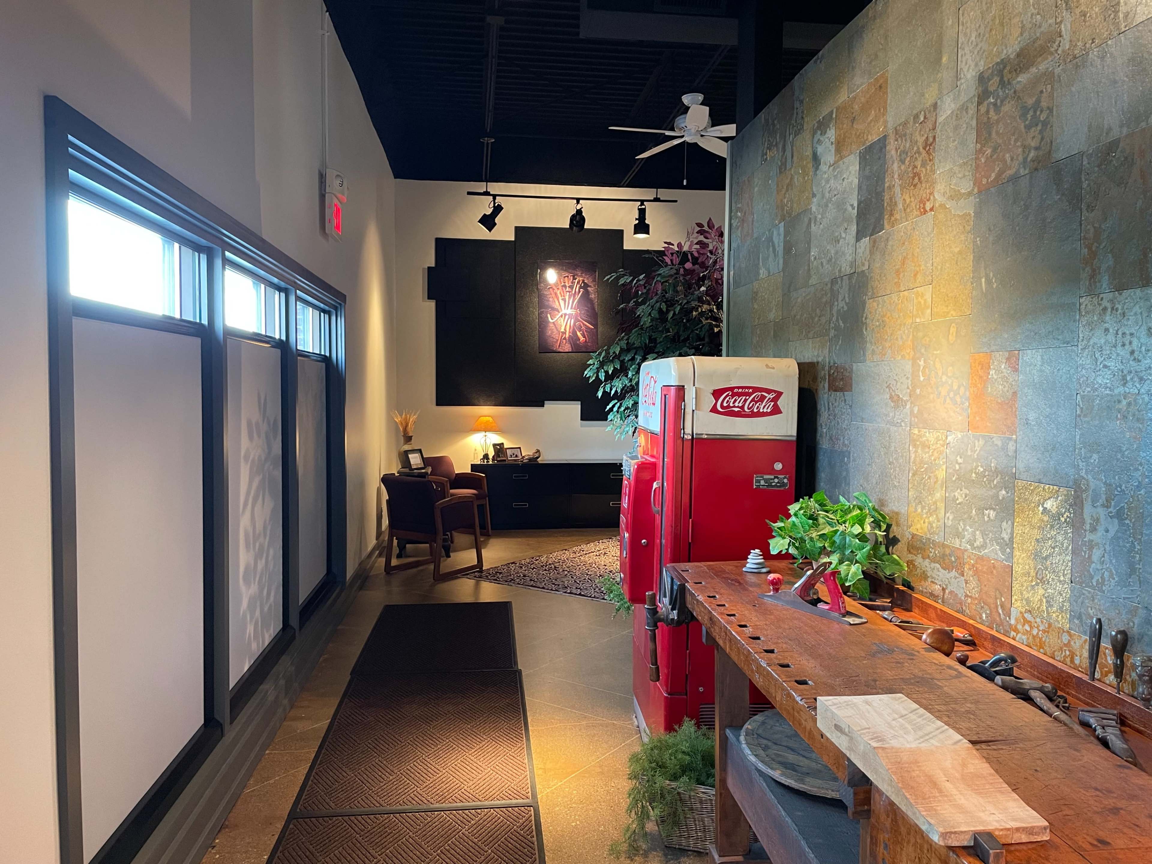 A hallway featuring a vintage Coca-Cola vending machine, a wooden workbench, and a desk area illuminated by soft lighting at the end.