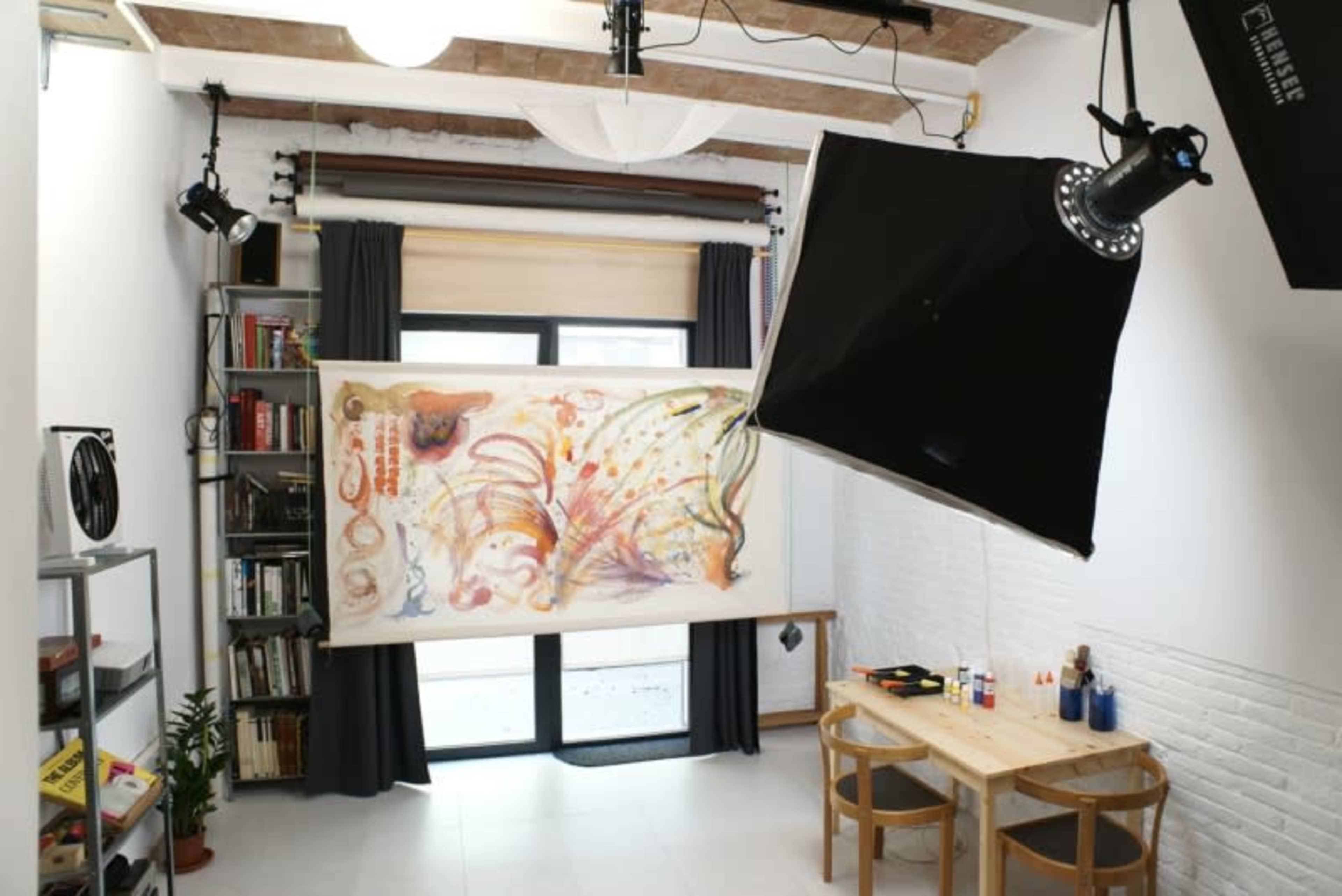 A well-lit studio features a large artwork hanging between two black curtains, with a wooden table and backdrop lighting equipment in the foreground.