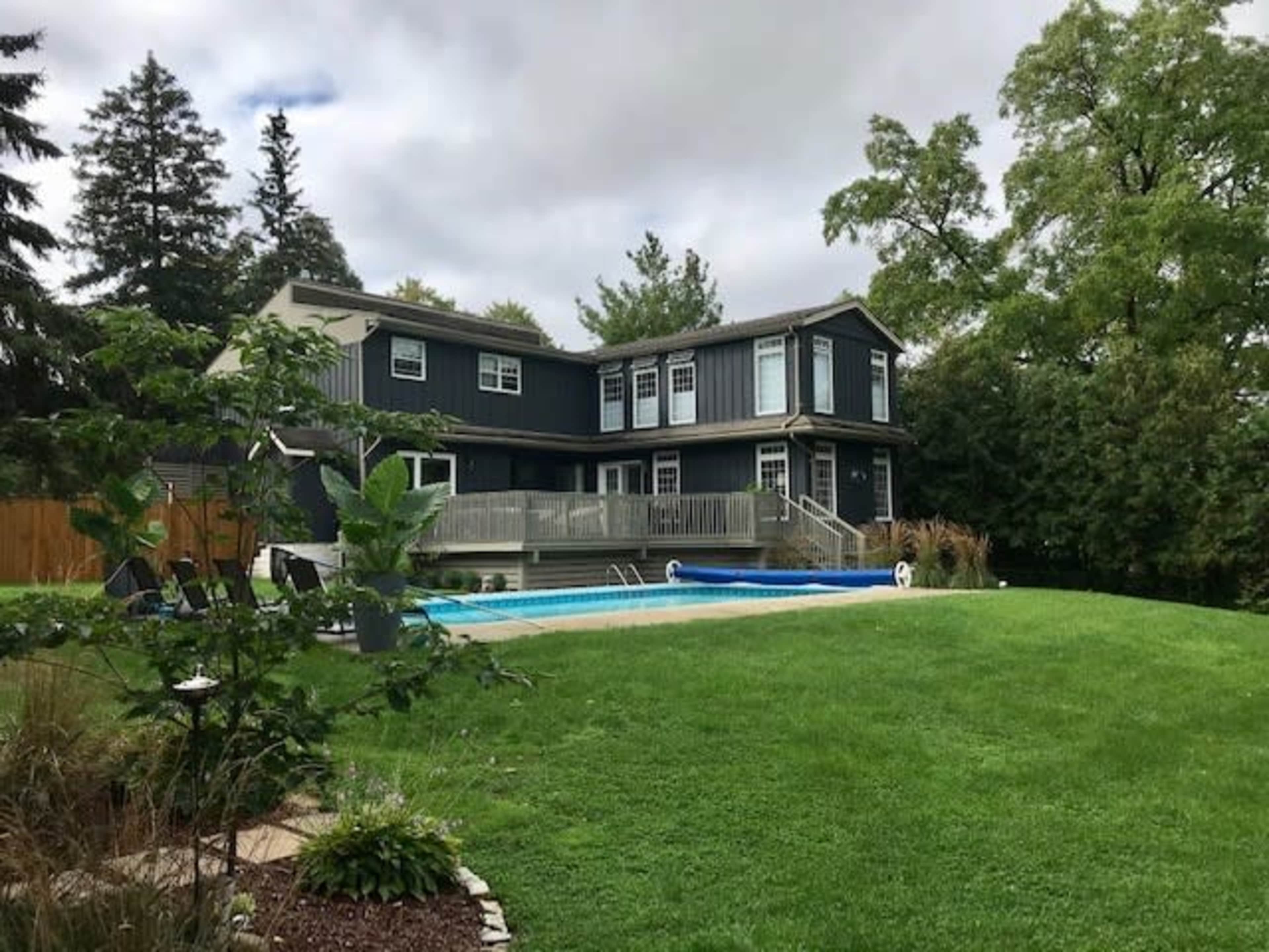A modern two-story house with a deck overlooks a swimming pool and landscaped lawn surrounded by trees.