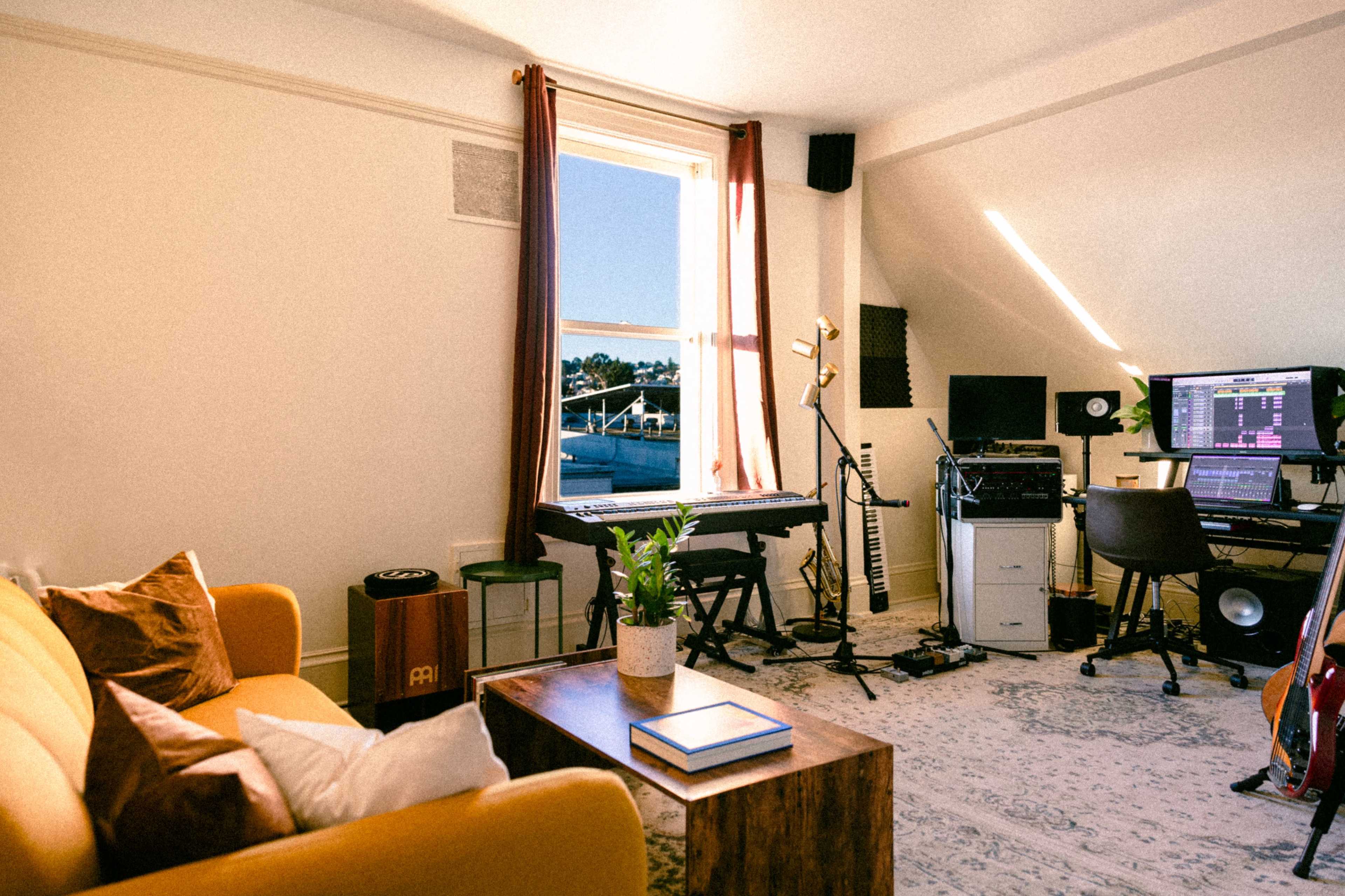 The scene shows a cozy room featuring a yellow sofa, a wooden coffee table, a keyboard and microphone set up, and a computer workstation with music equipment, illuminated by natural light coming through a window.