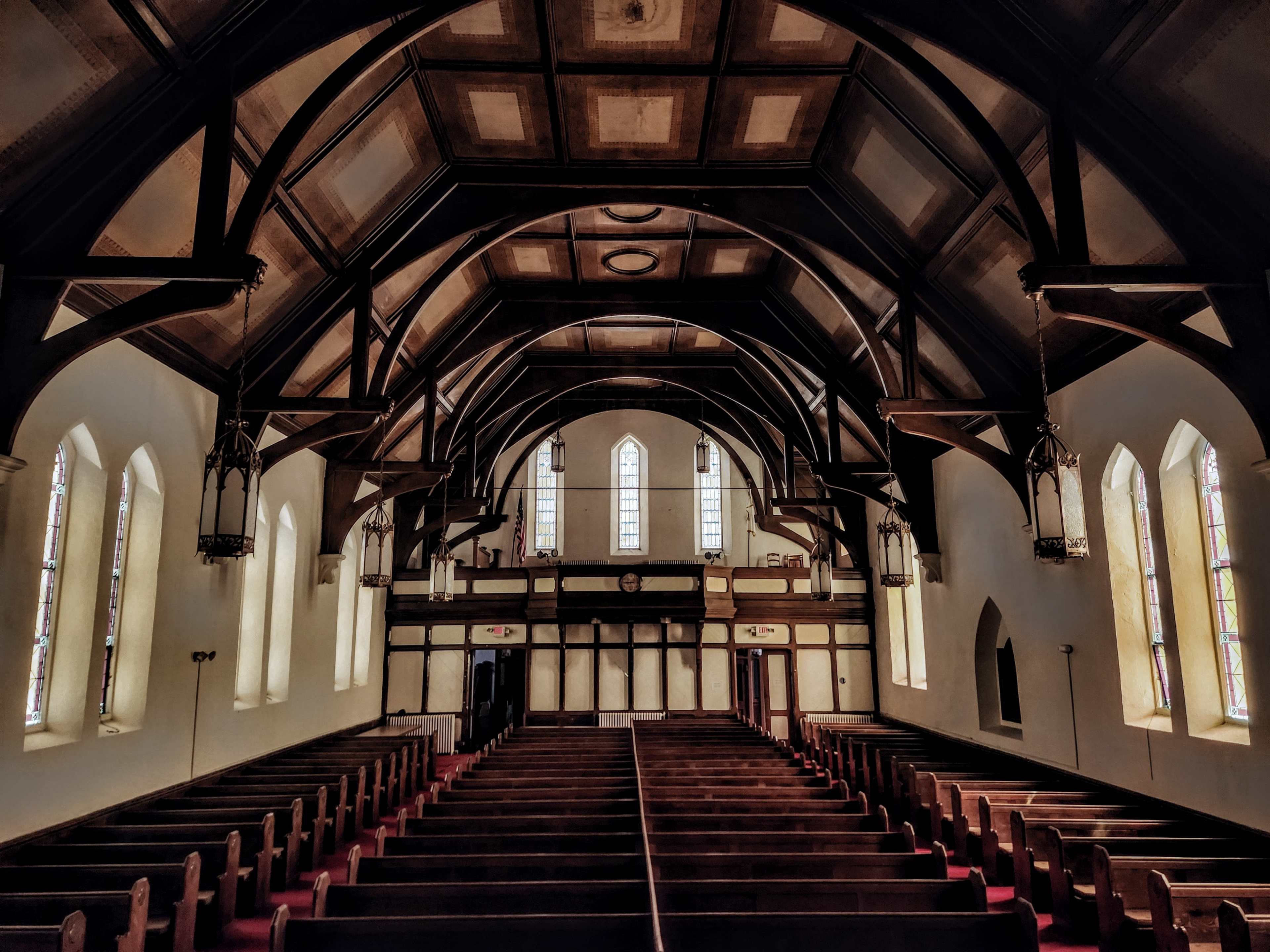 The interior of an empty church features wooden pews arranged in rows beneath a high vaulted ceiling with decorative beams and stained glass windows.