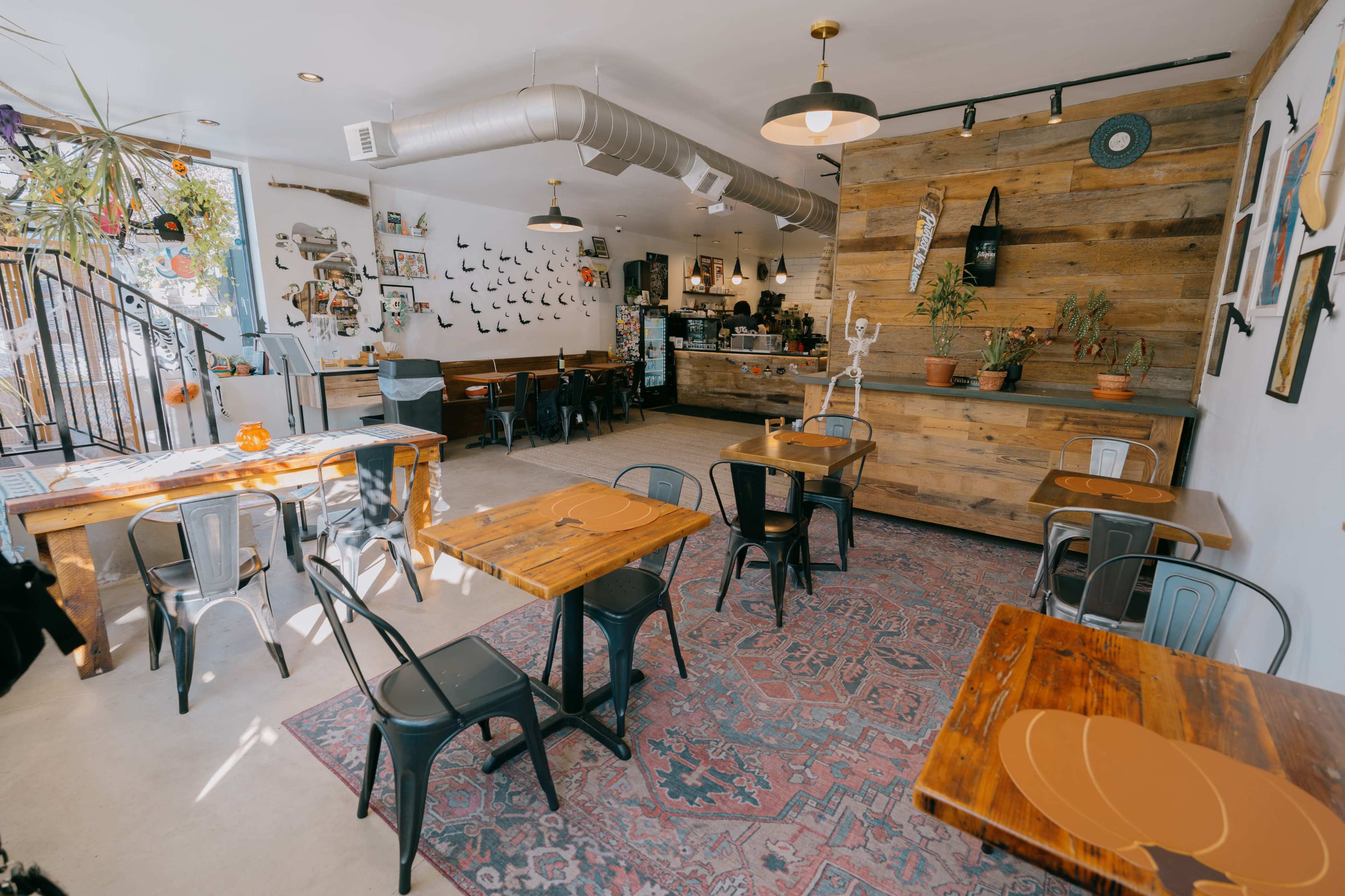 The image shows a cozy café interior featuring wooden tables, metal chairs, decorative plants, and various artworks on the walls.