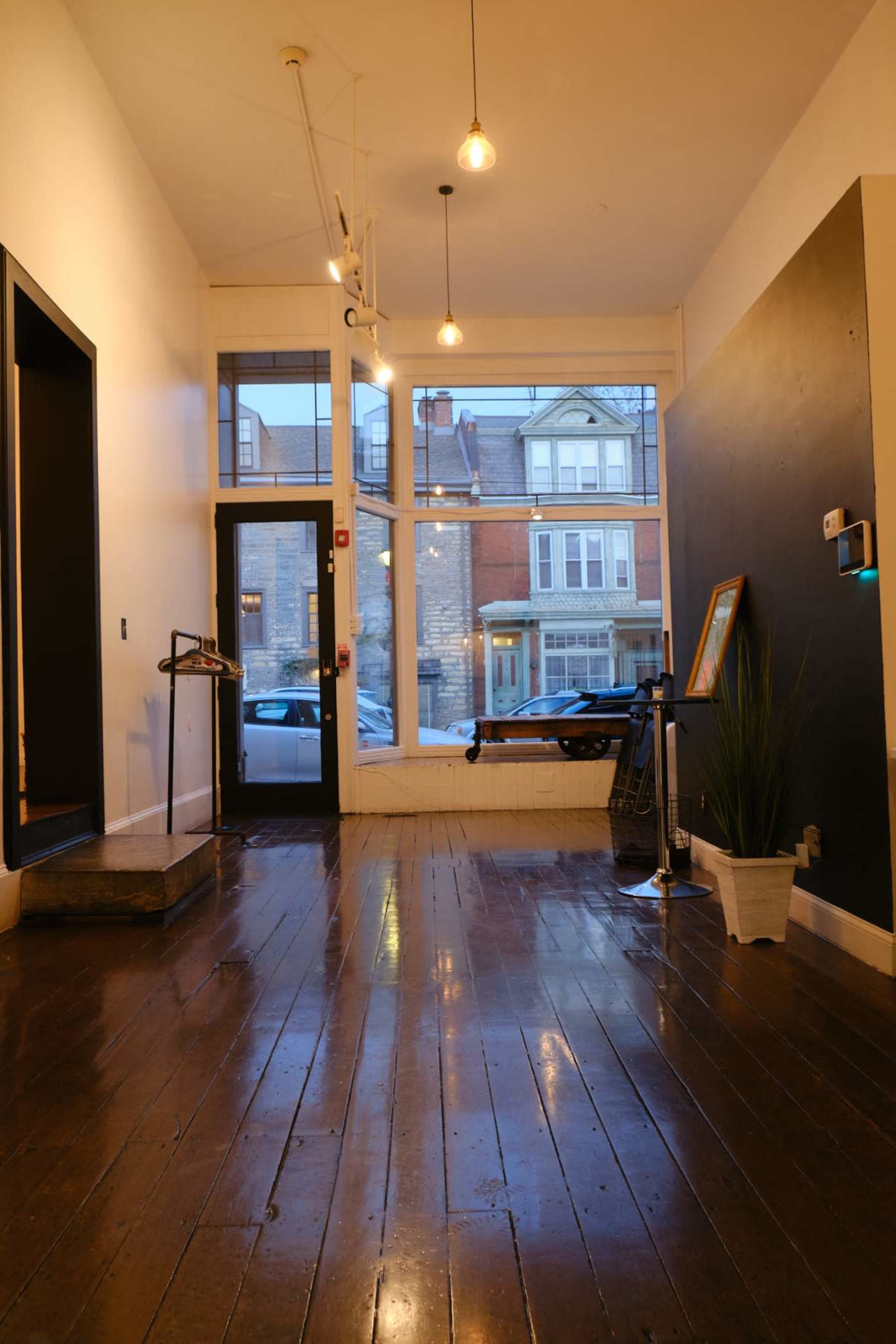 The image shows a spacious, empty room with polished wooden floors, large front windows, and a black accent wall.