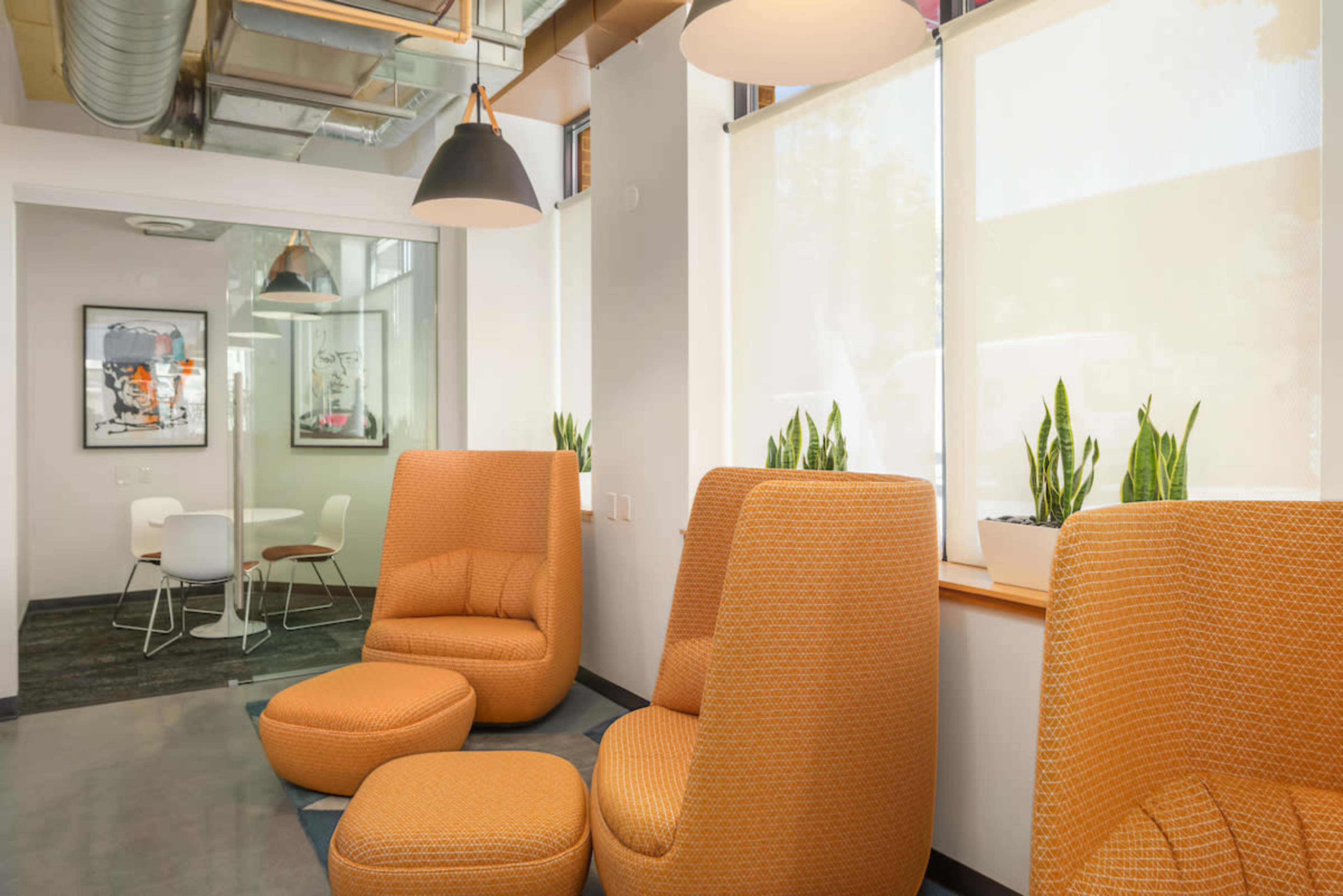 A modern interior space featuring two orange lounge chairs and a round ottoman, with a small table and chairs visible in the background.