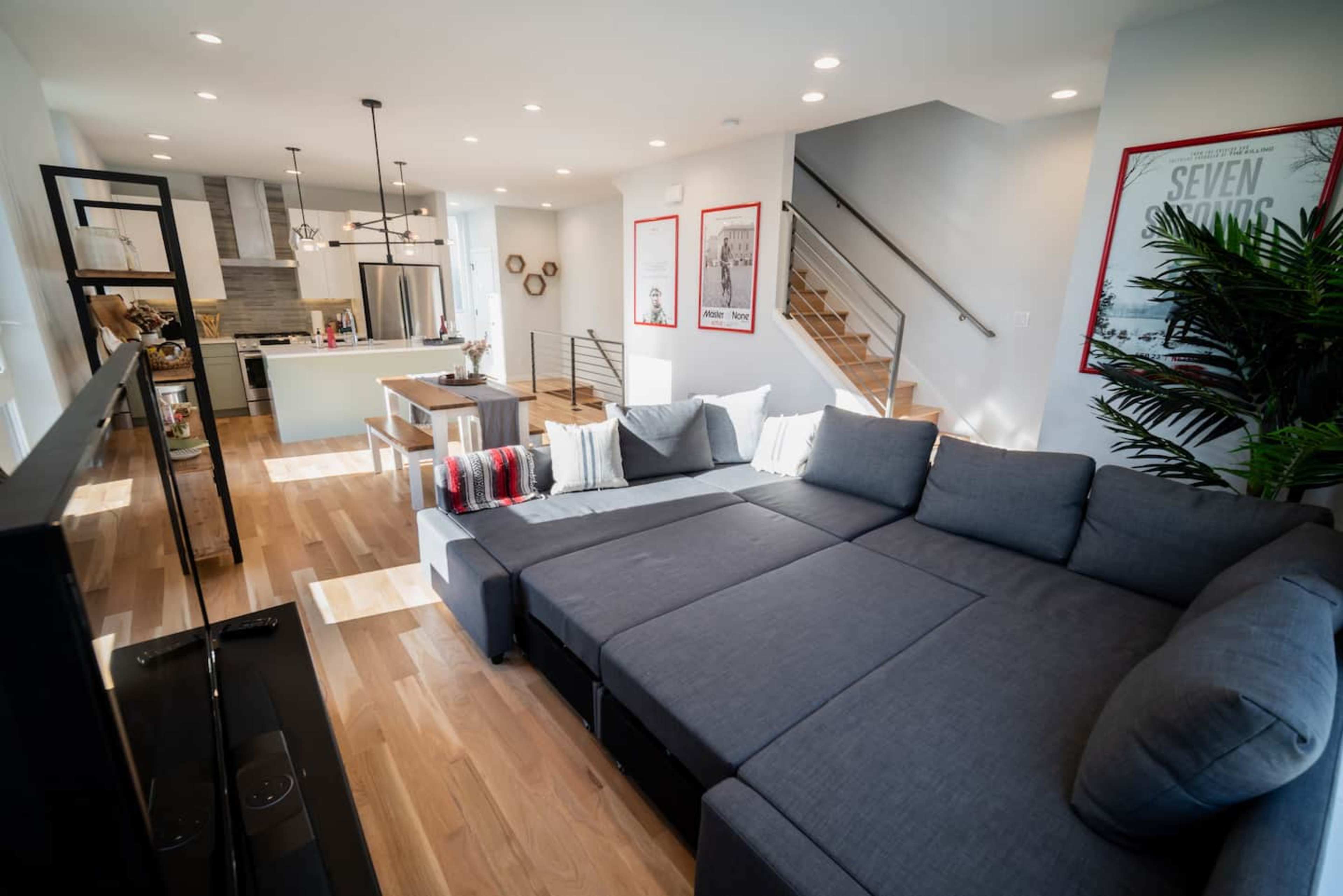 A modern living room with a gray sectional sofa, a kitchen in the background, and a staircase leading to the upper floor.