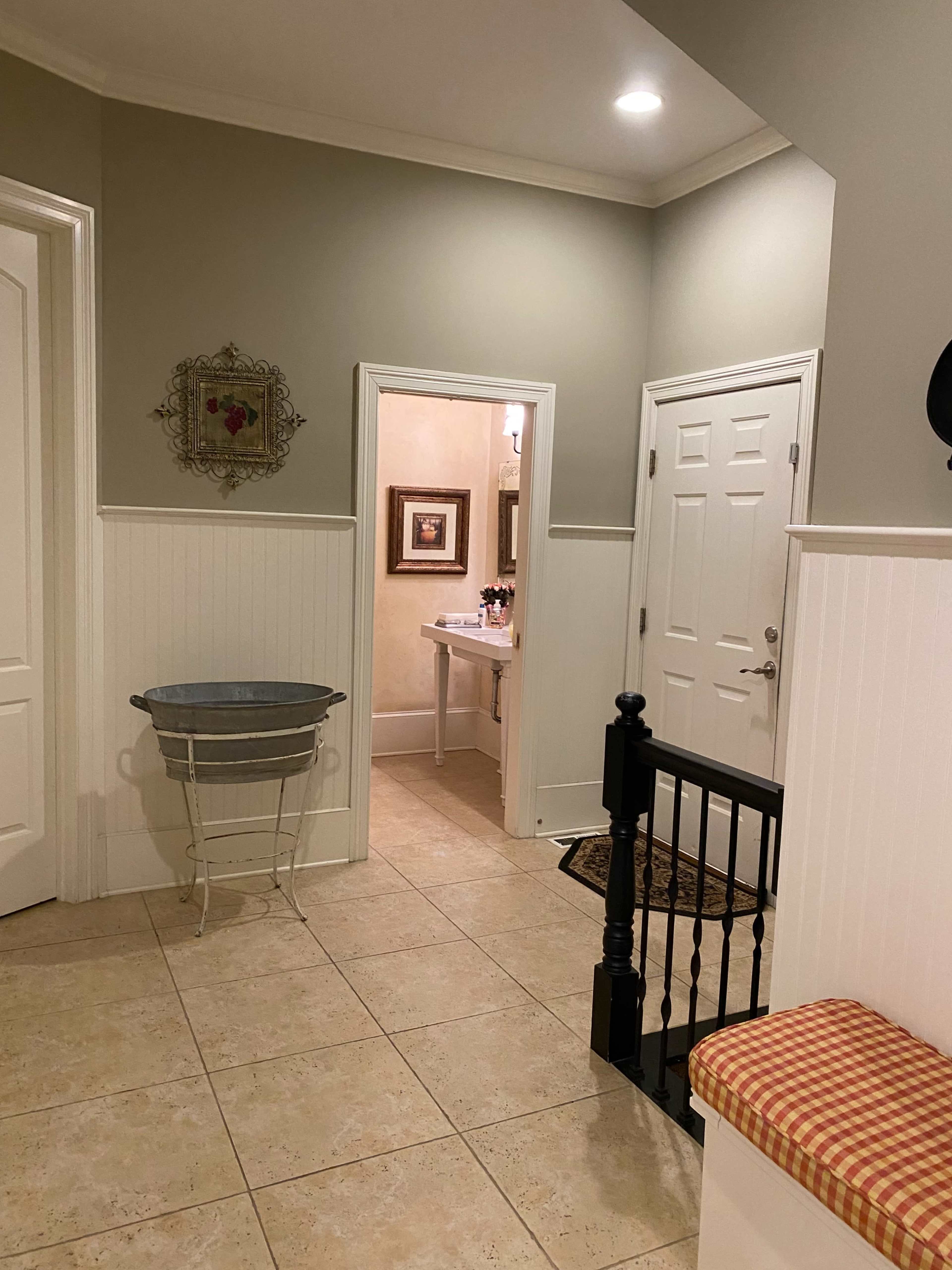 A hallway with beige tiled flooring, white wainscoting, and a view of a bathroom at the end.