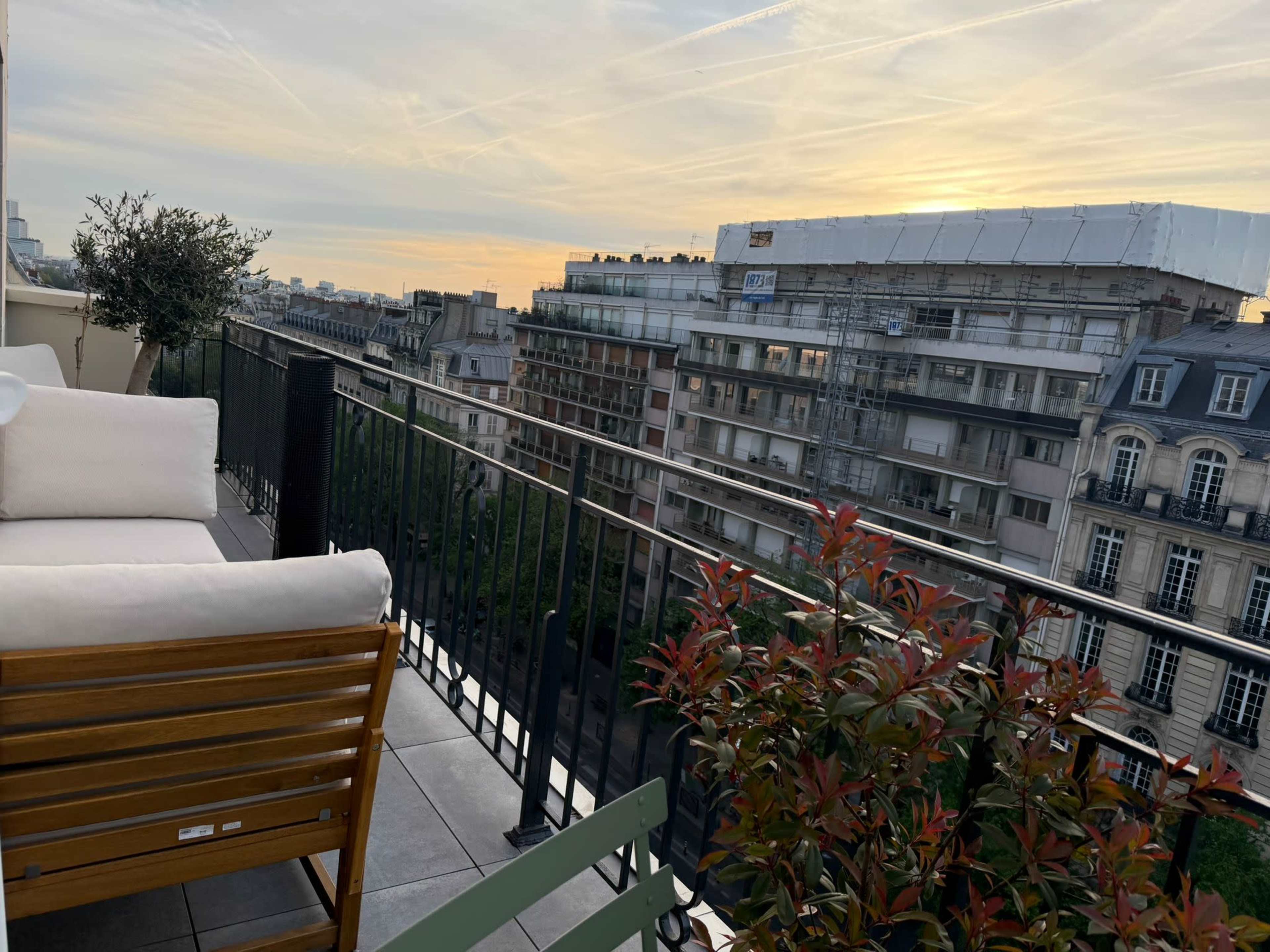 The image shows a balcony with a wooden chair and potted plants, overlooking a cityscape at sunset.