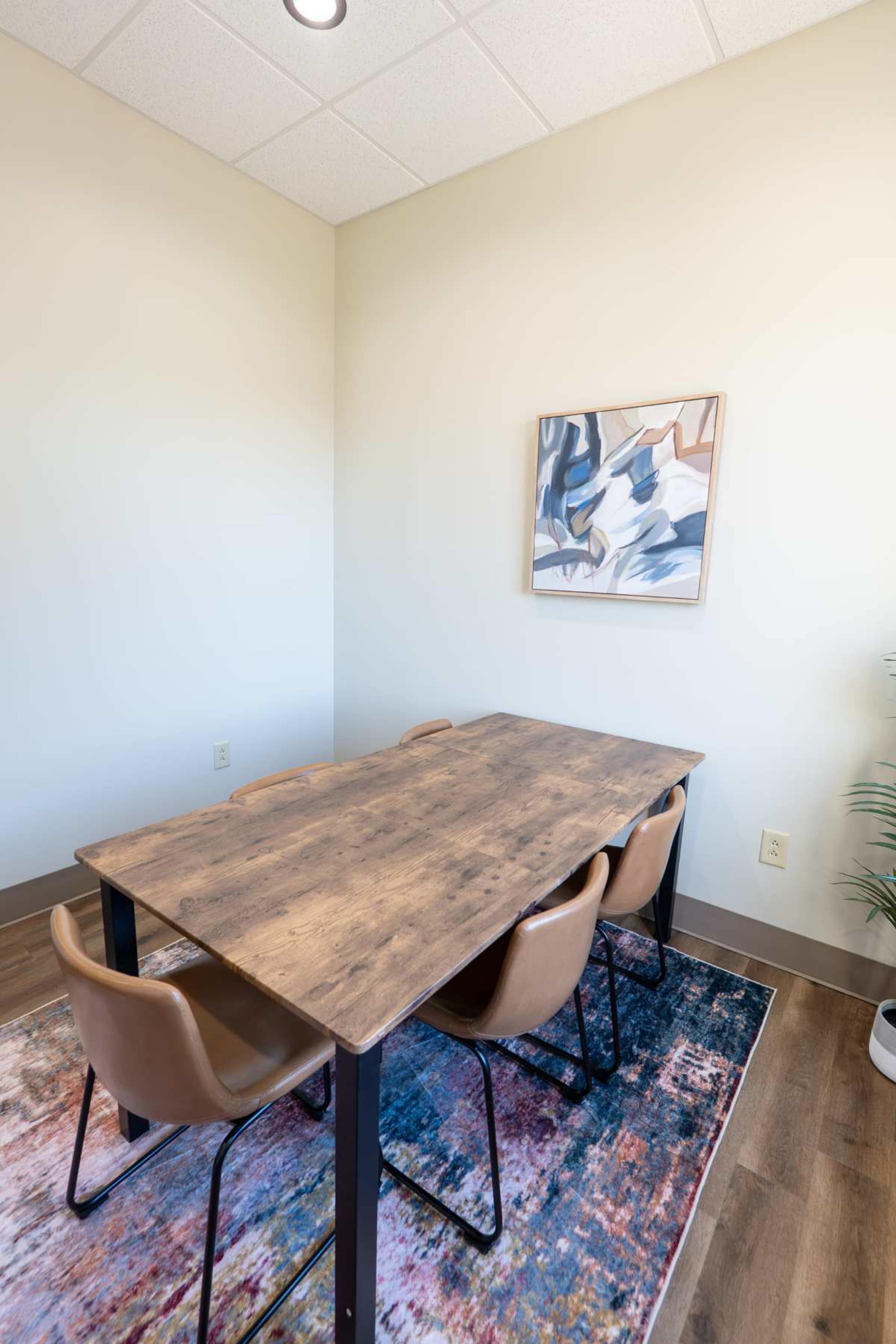 The image shows a small meeting room with a wooden table surrounded by four brown chairs, a colorful rug on the floor, and a piece of abstract art hung on the wall.