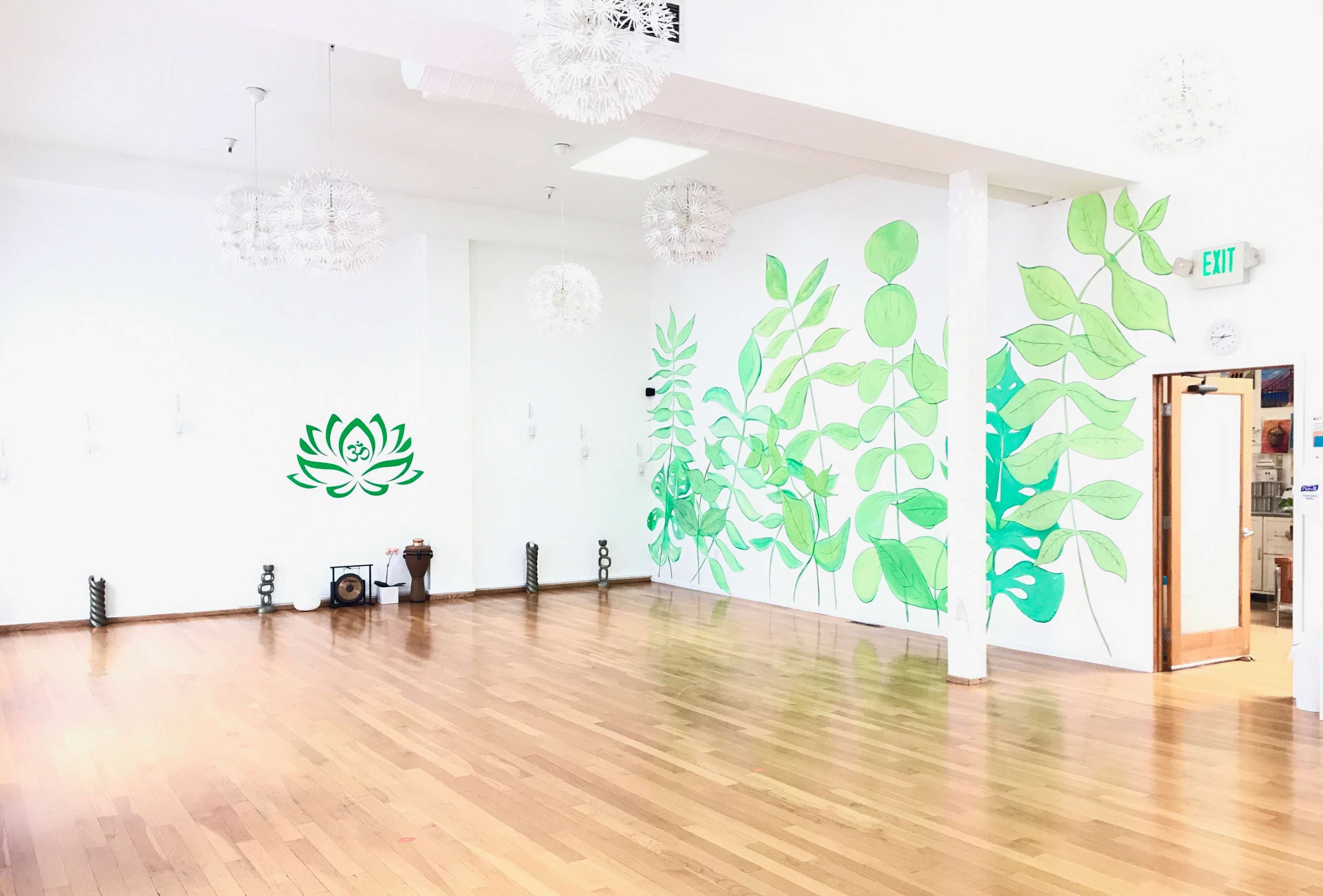 A spacious, bright room with a polished wooden floor, featuring large green leaf murals on one wall and decorative light fixtures hanging from the ceiling.