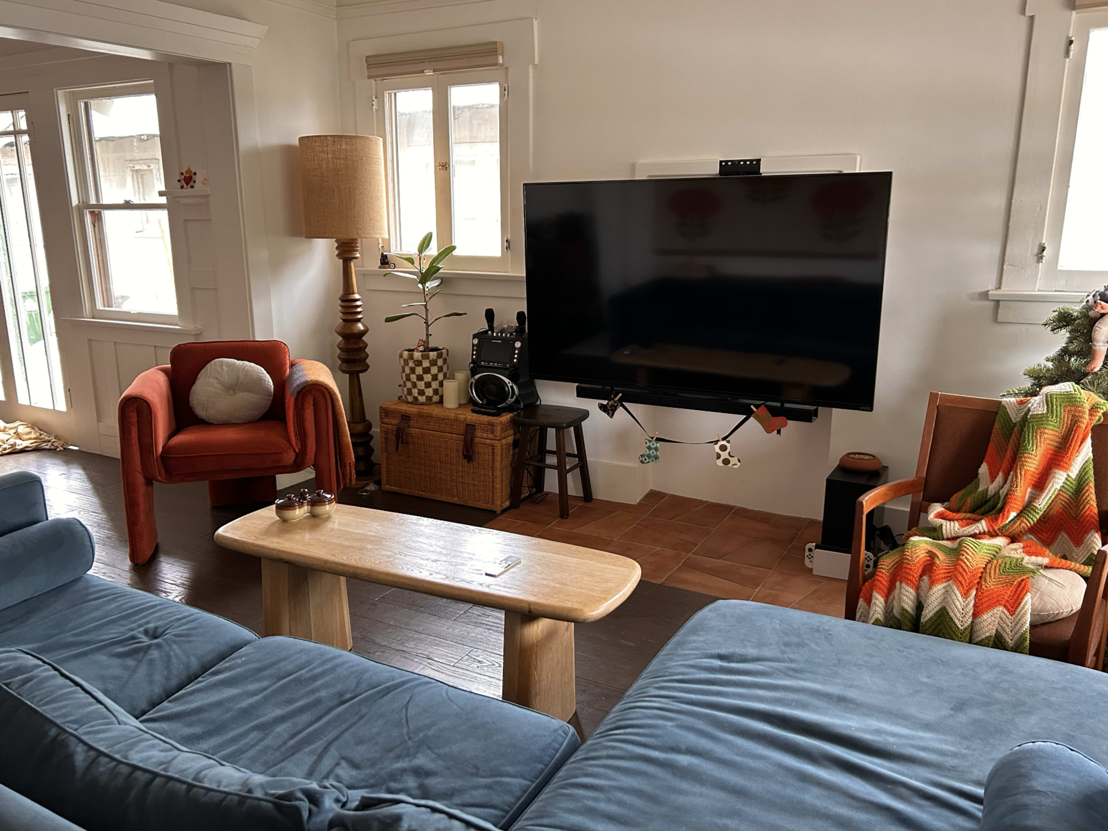 The image depicts a cozy living room with blue couches, a wooden coffee table, a mounted television, and decorative elements including a lamp and a colorful blanket.