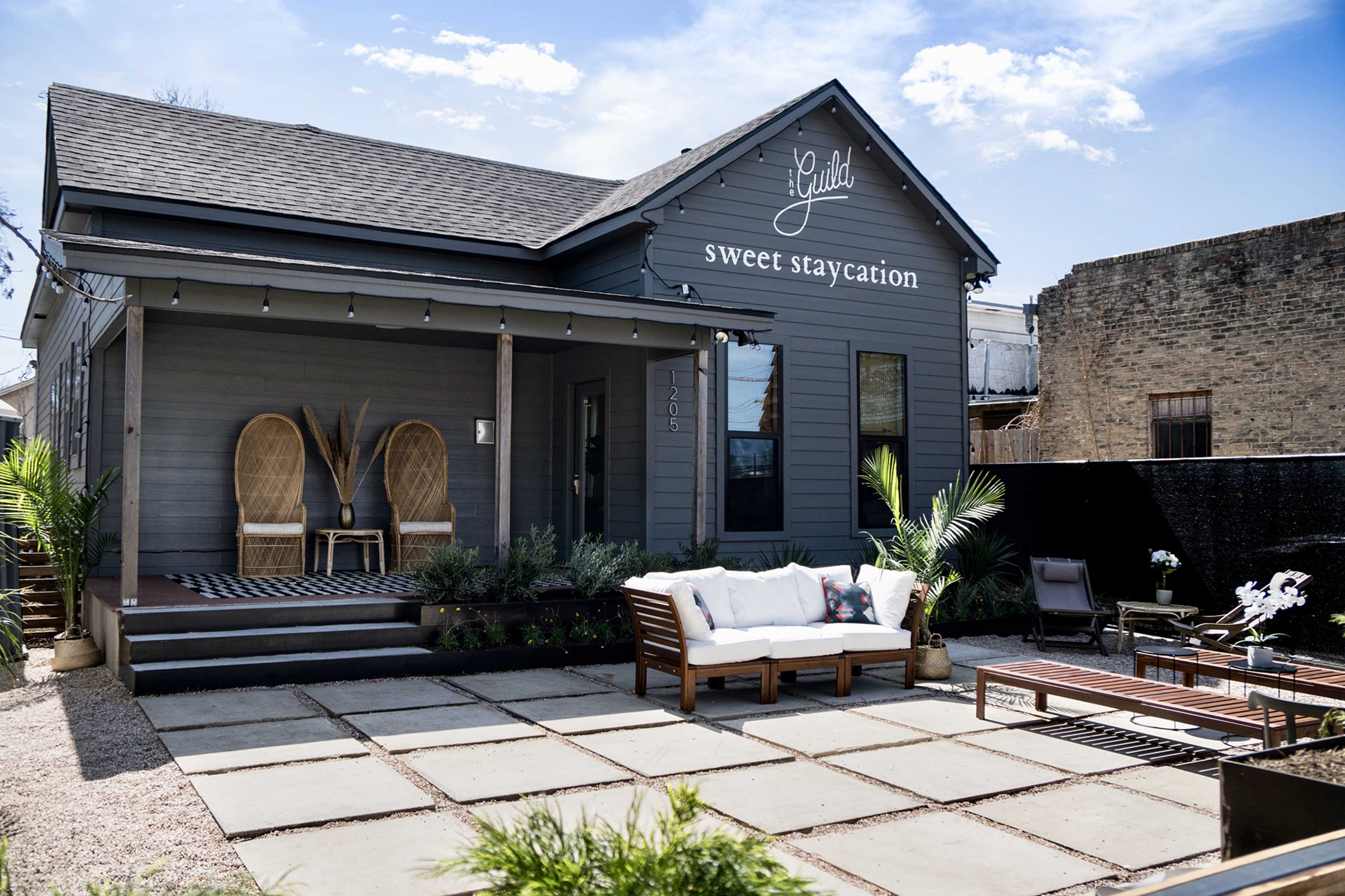 A gray house with a sign reading "sweet staycation" features a front porch with two wicker chairs and a seating area made of stone pavers.