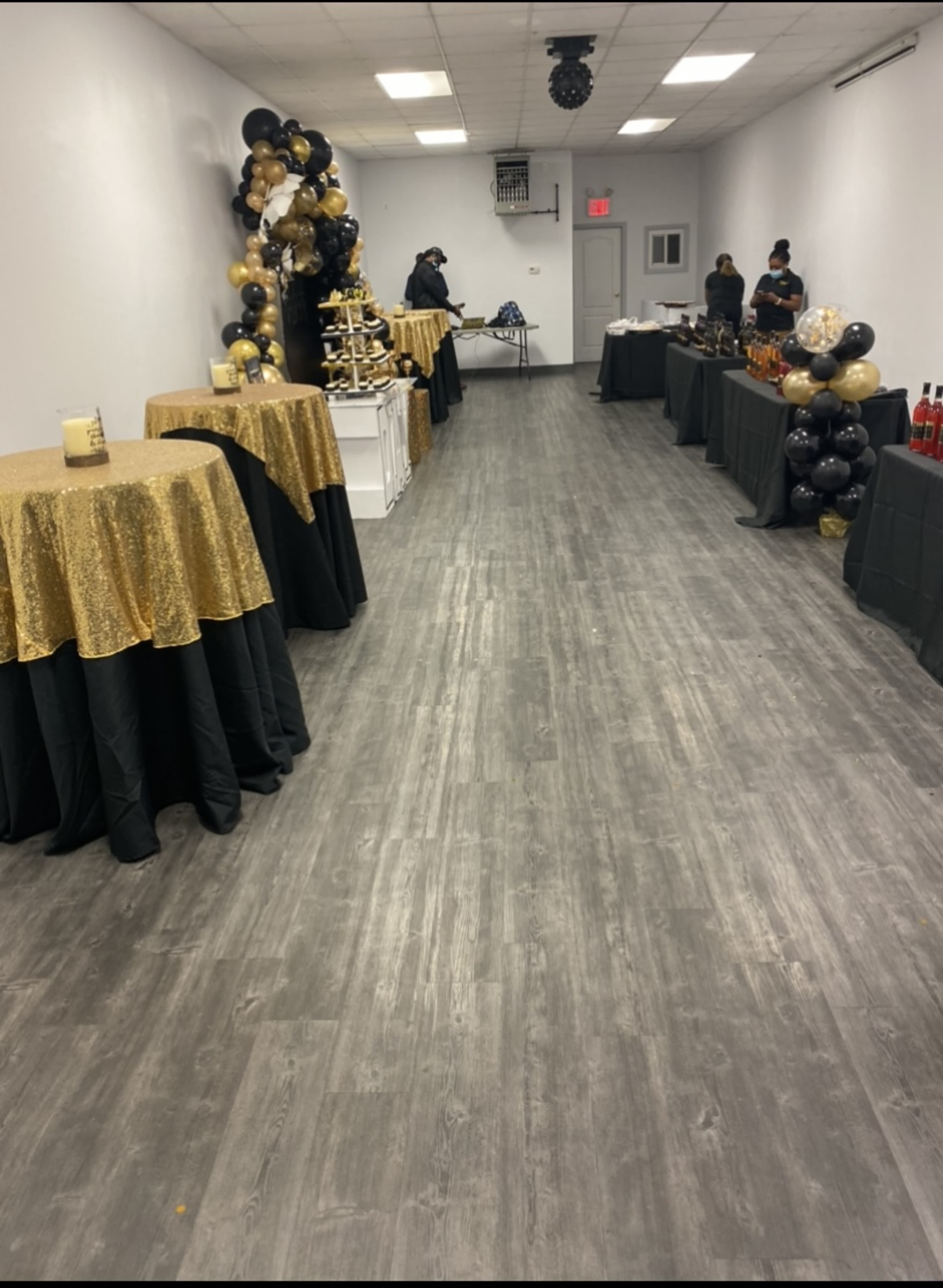 A spacious, decorated room with tables covered in gold and black cloths, set up for an event, along with a display of drinks on tables at either end.