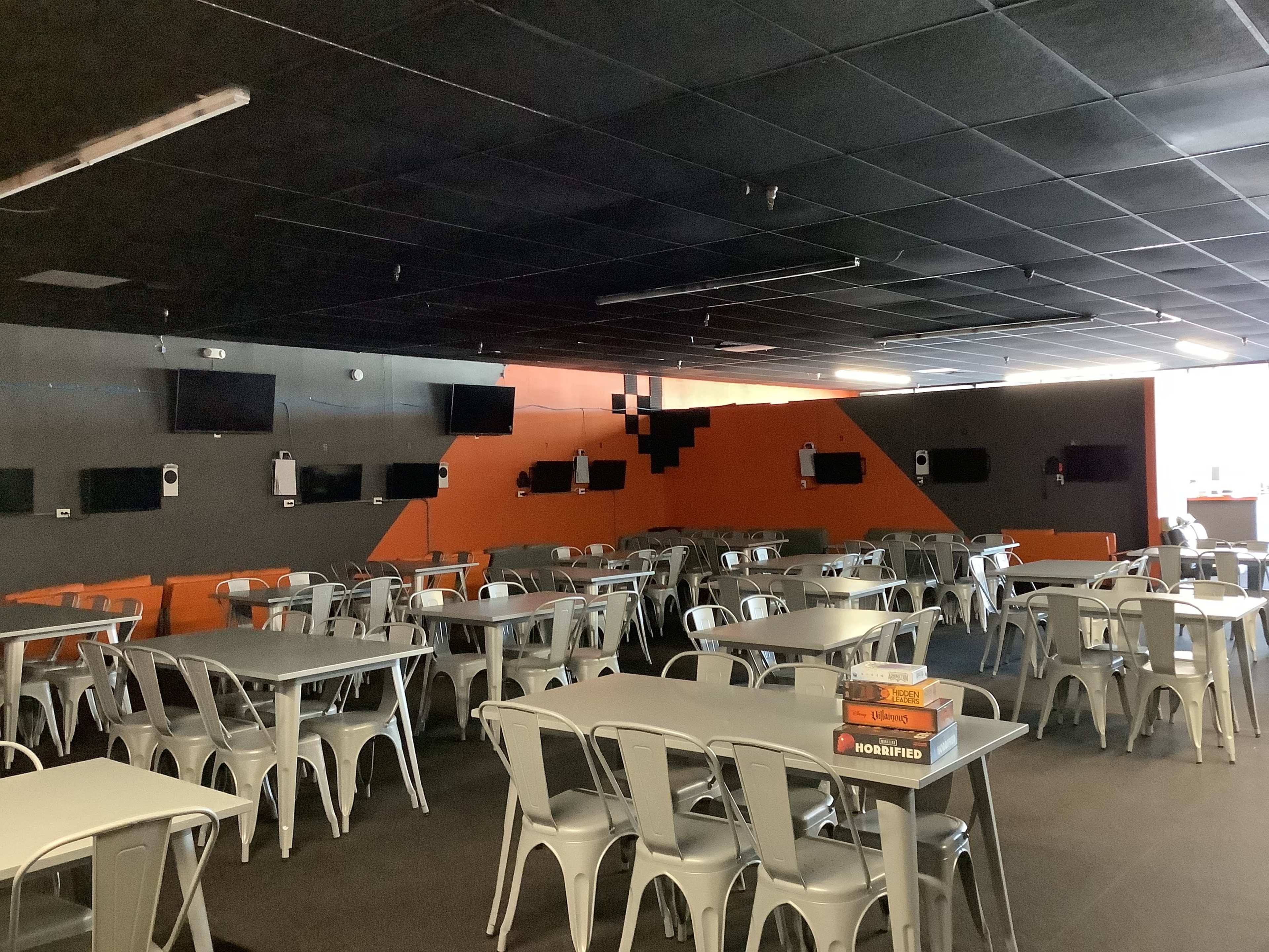 A spacious indoor area features metal tables and chairs arranged neatly, with several wall-mounted screens and a vibrant orange accent wall.