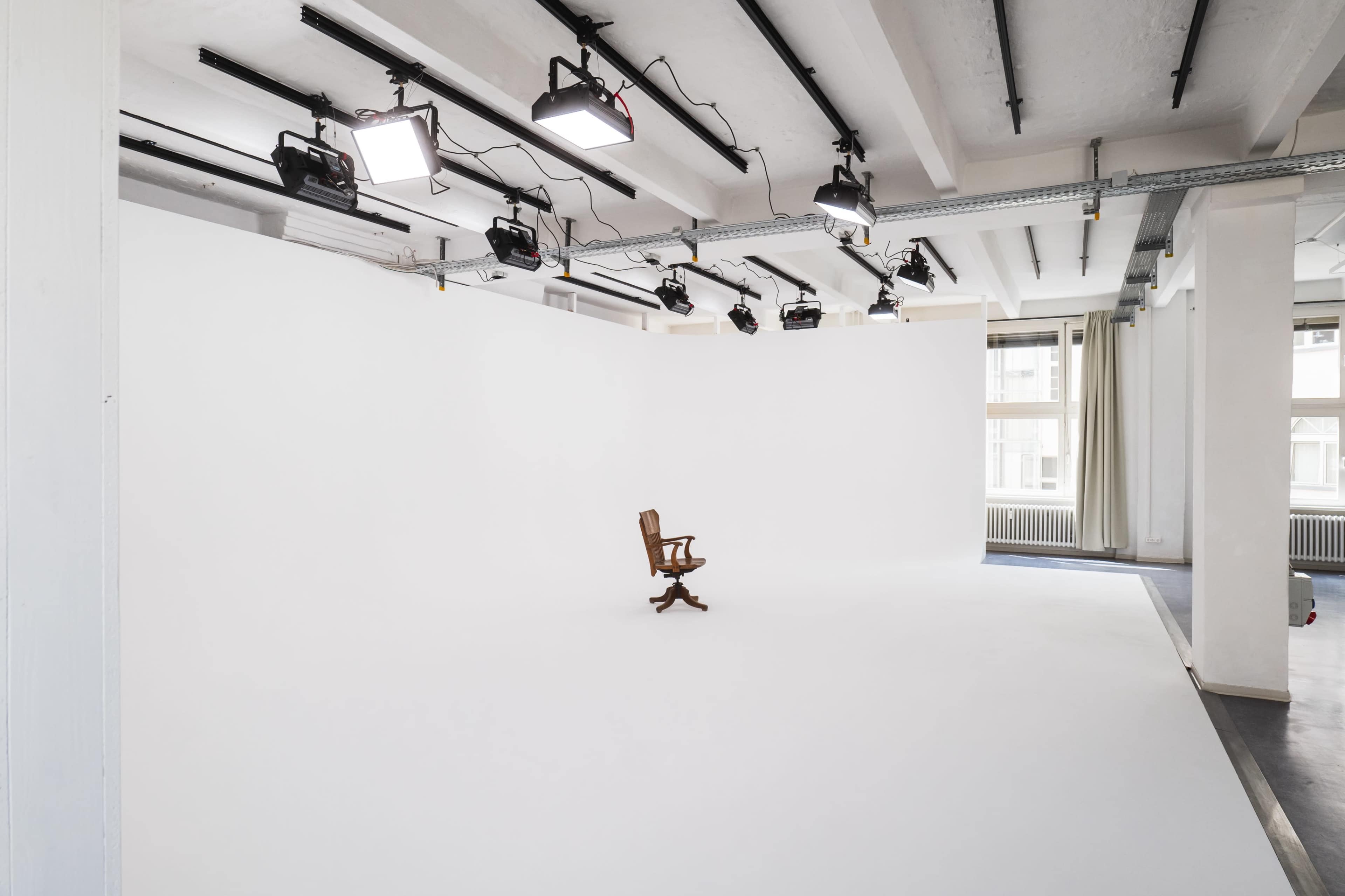 A single wooden chair is positioned in the center of a spacious, bright studio with white walls and overhead lighting.