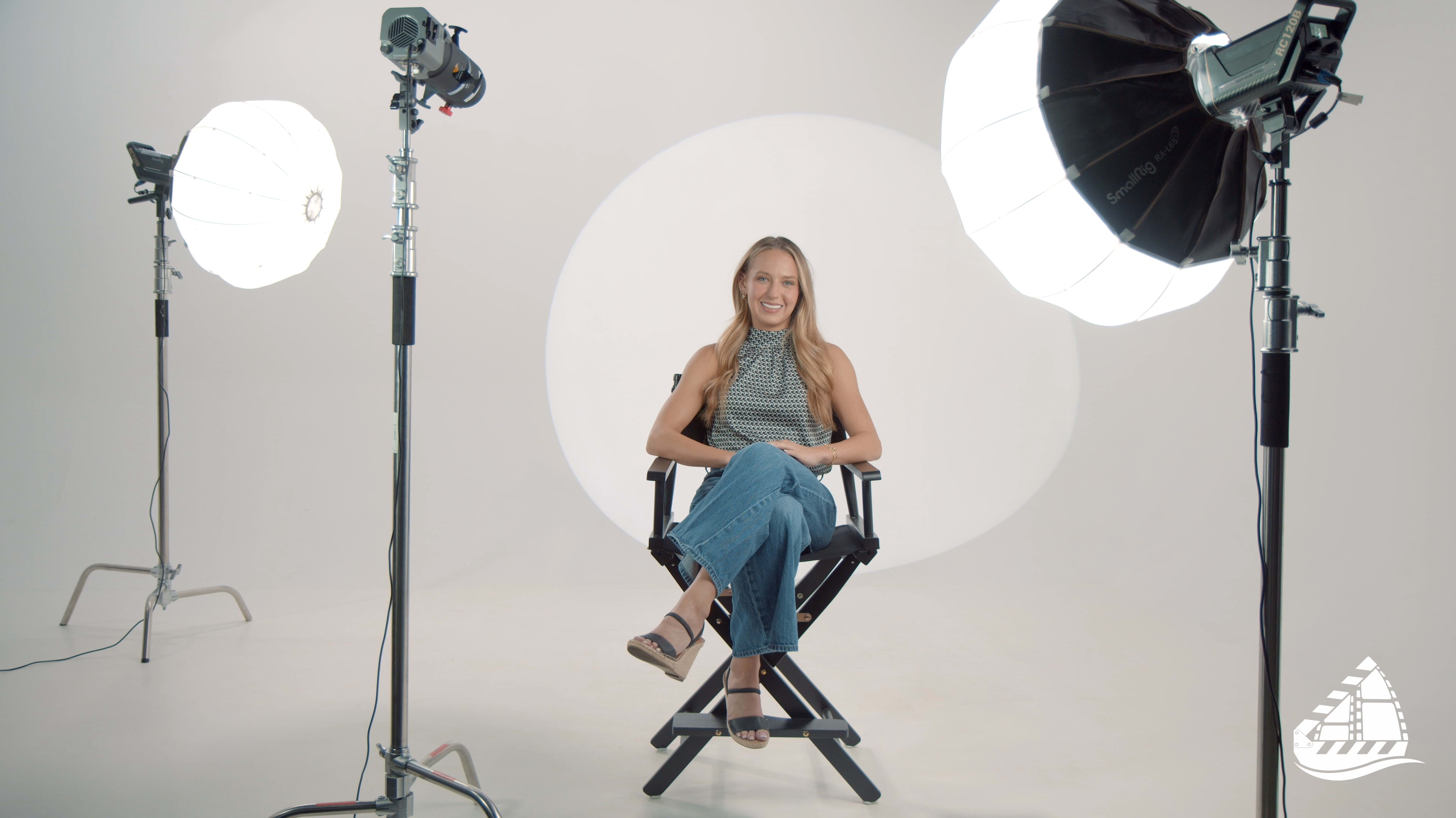 A smiling woman sits on a director's chair in a studio setting, surrounded by softbox lighting equipment.