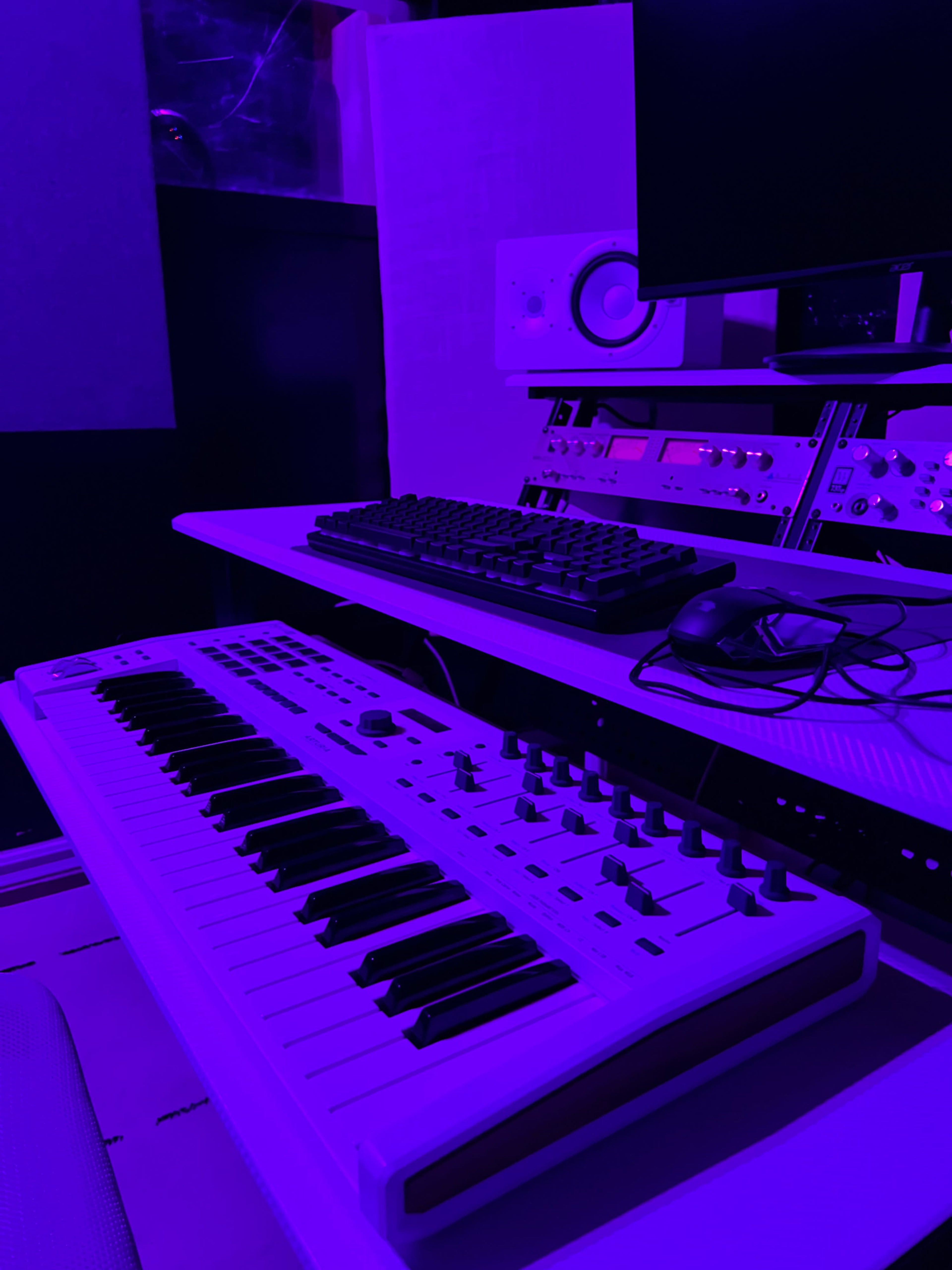 A keyboard and a computer keyboard are positioned on a desk in a dimly lit music studio with purple lighting.
