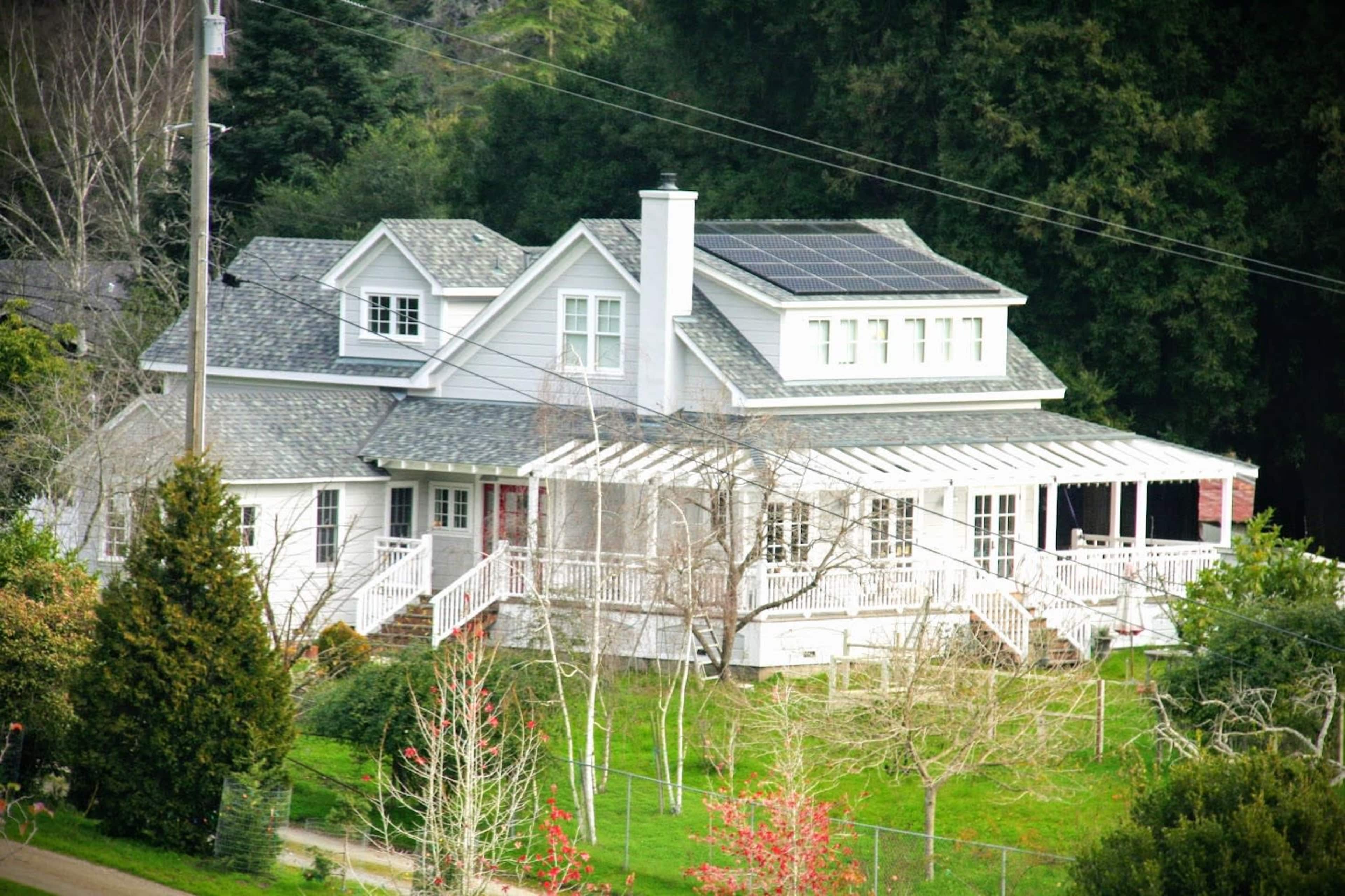 A two-story house with a gabled roof and solar panels is surrounded by trees and has a front porch with a railing.