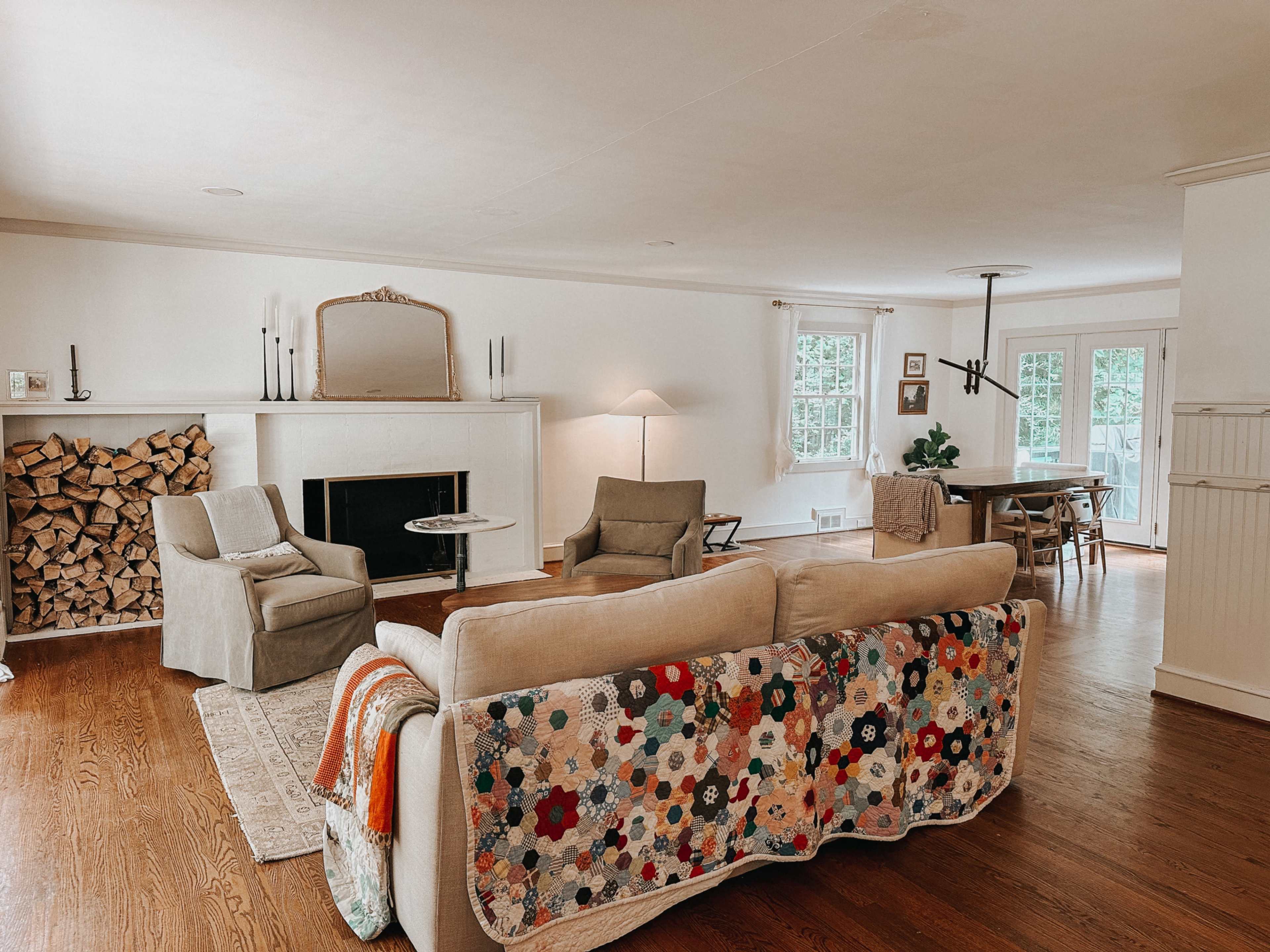 A living room with a sofa covered in a colorful quilt, two chairs, a fireplace with stacked wood, and a dining area in the background.