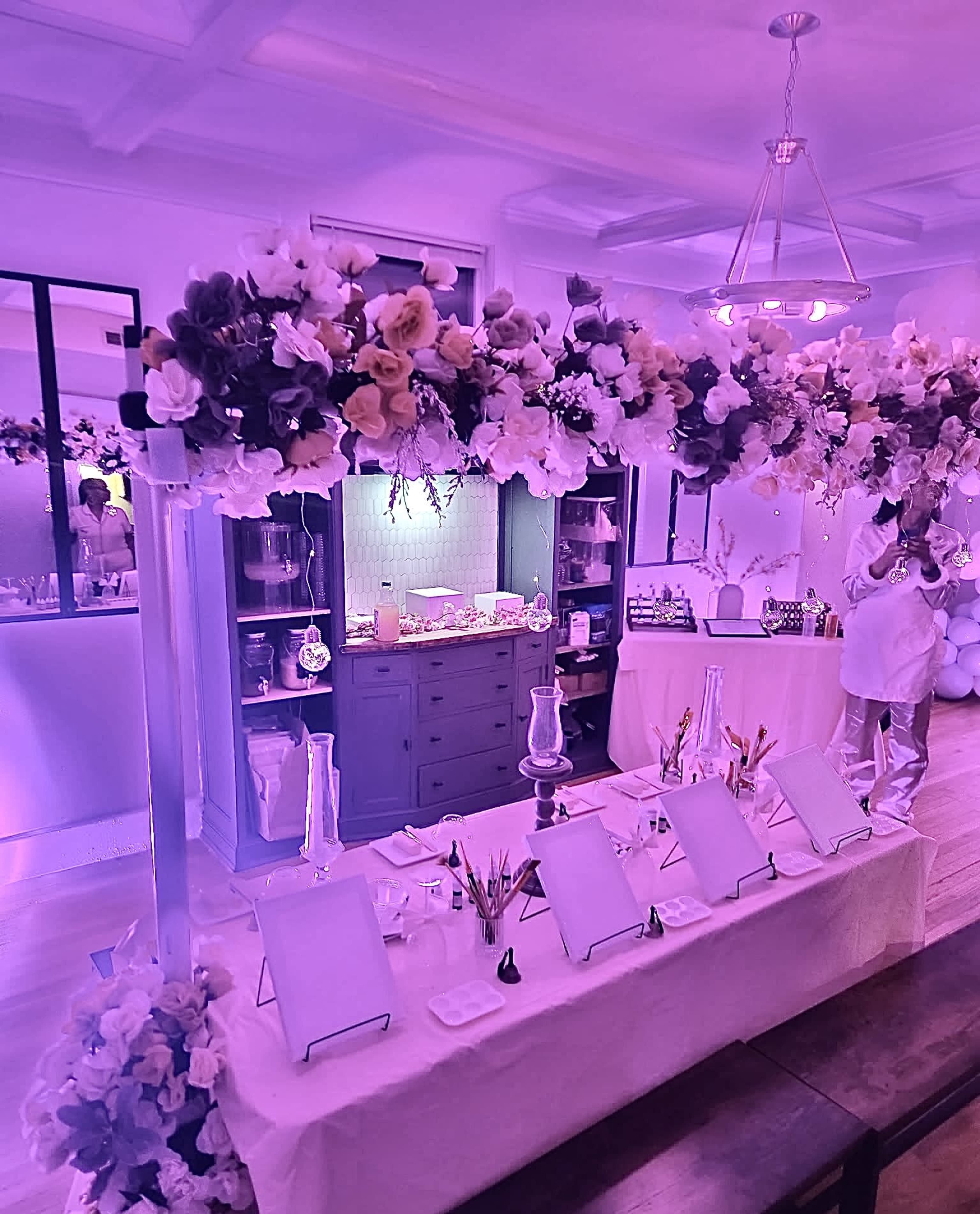 A decorated event space features a floral arrangement above a service area with empty display frames and party supplies, illuminated by purple lighting.