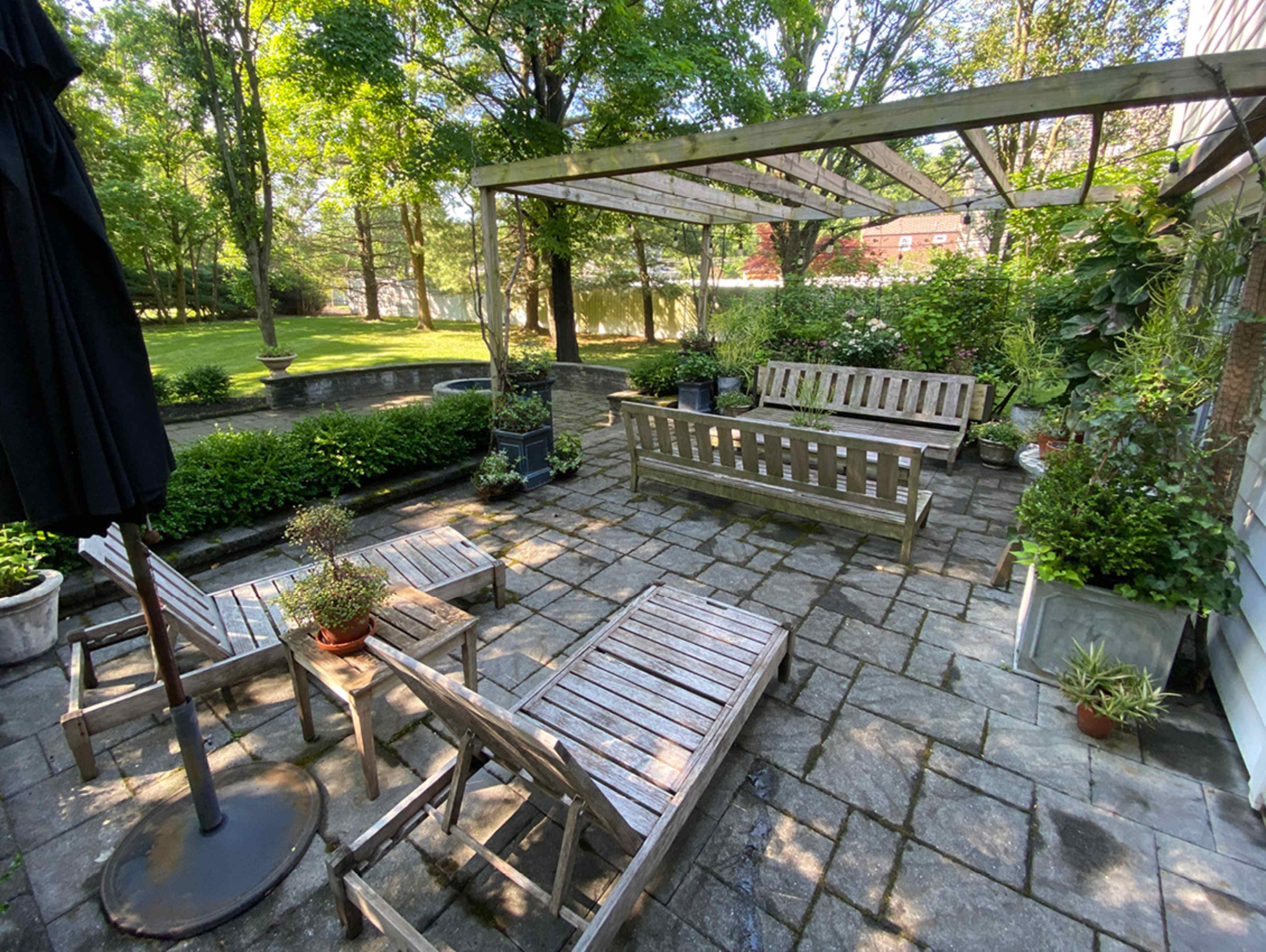 A stone patio features wooden furniture and potted plants under a wooden pergola in a shaded outdoor space.