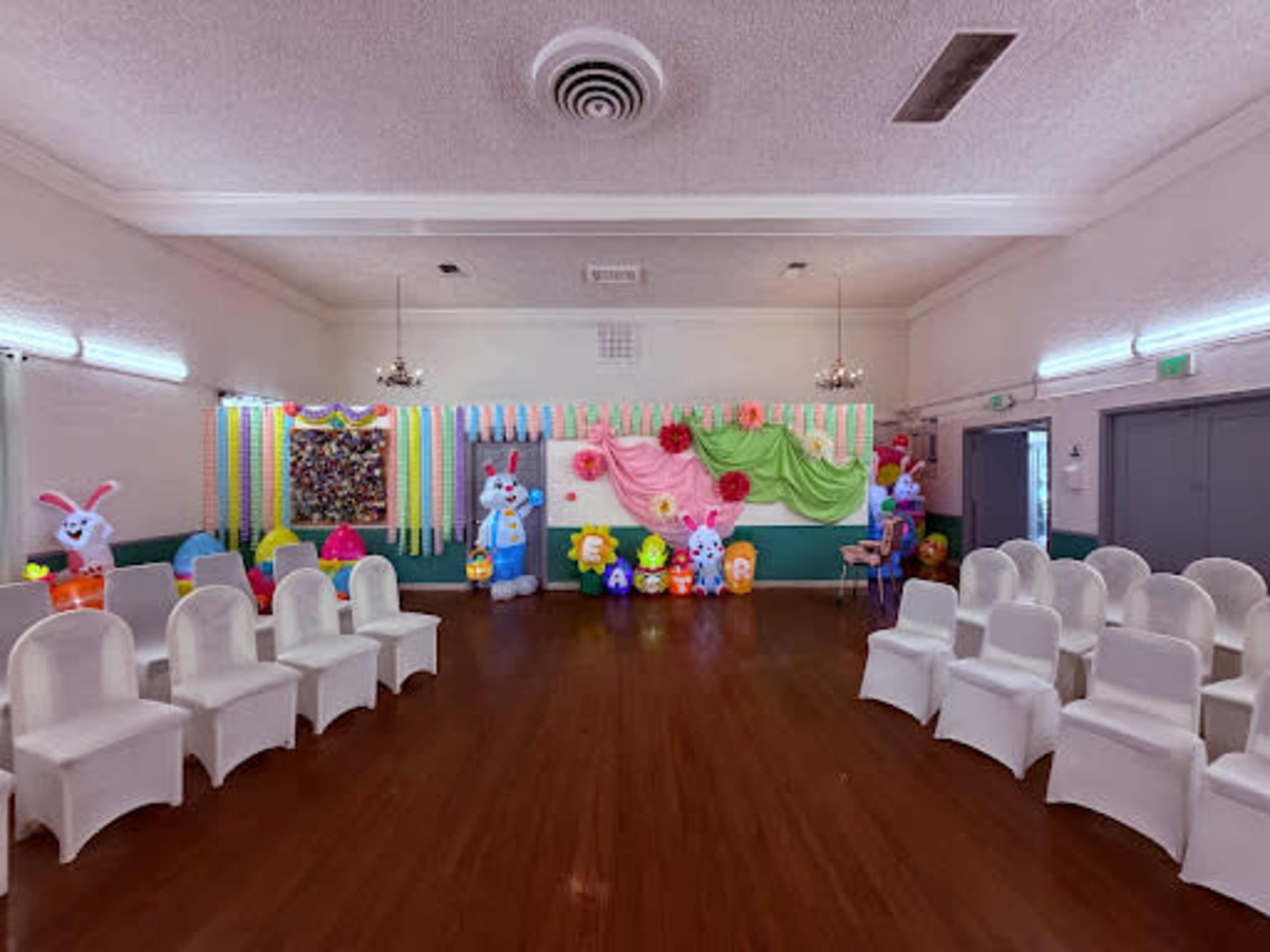 The image shows a decorated indoor venue with colorful streamers and balloons, along with a row of empty white chairs arranged facing a wall.