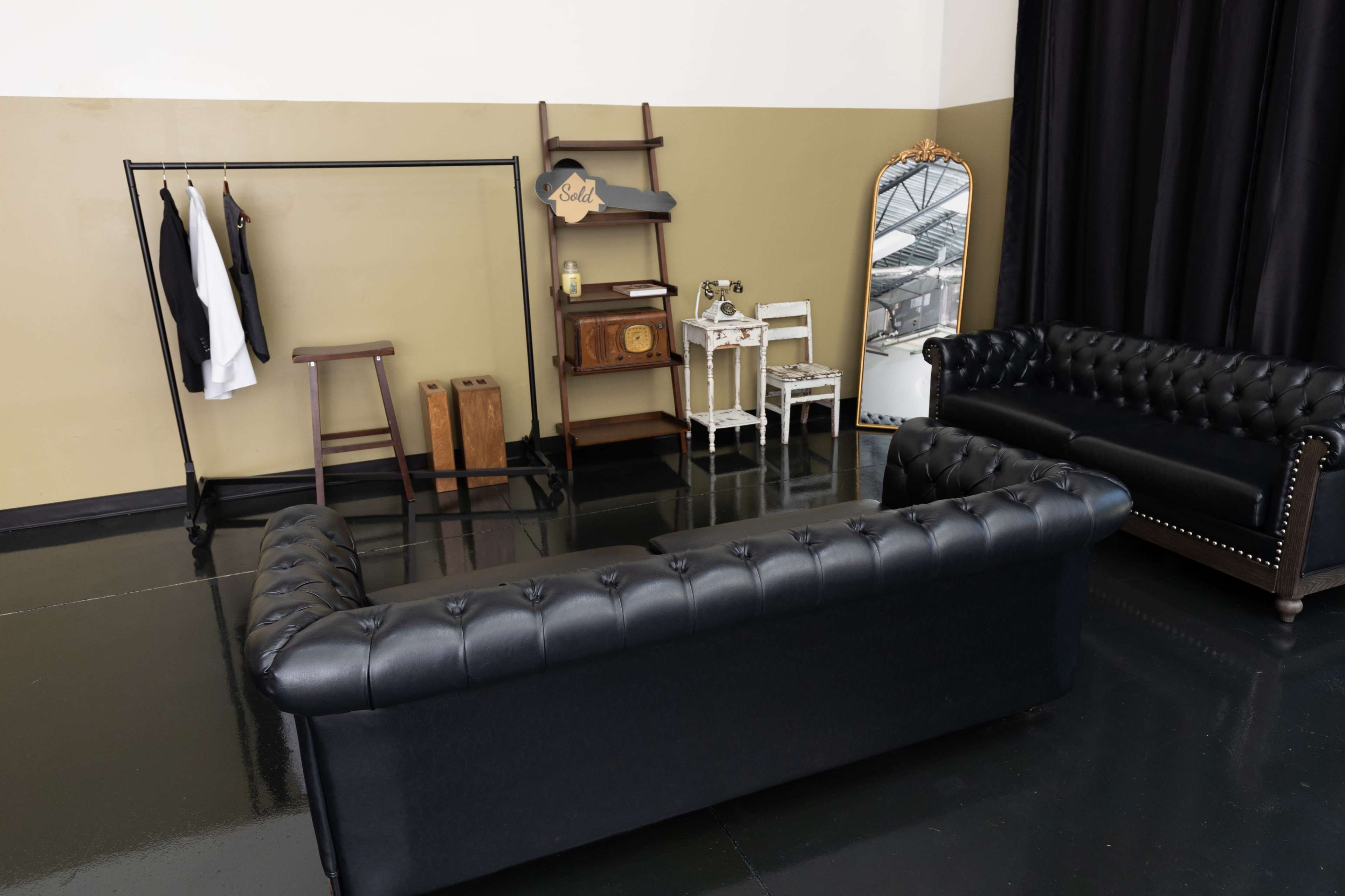 The image shows a room with a black leather chesterfield sofa, a clothing rack, a wooden ladder shelf, a small table with a vintage clock, and a mirror against a two-tone wall.