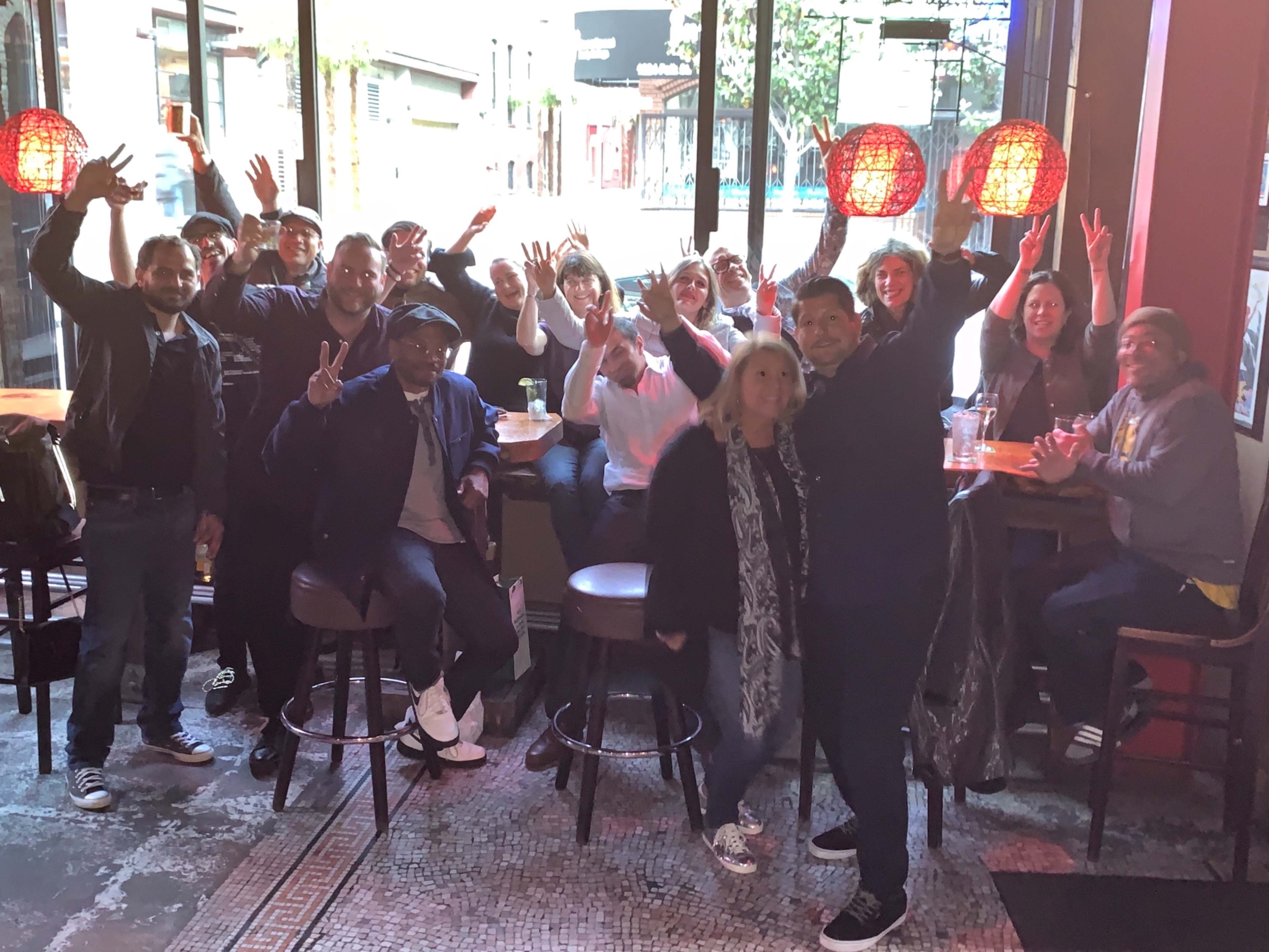 A group of people poses for a photo with their hands raised in a bar setting, surrounded by tall windows and hanging red light fixtures.