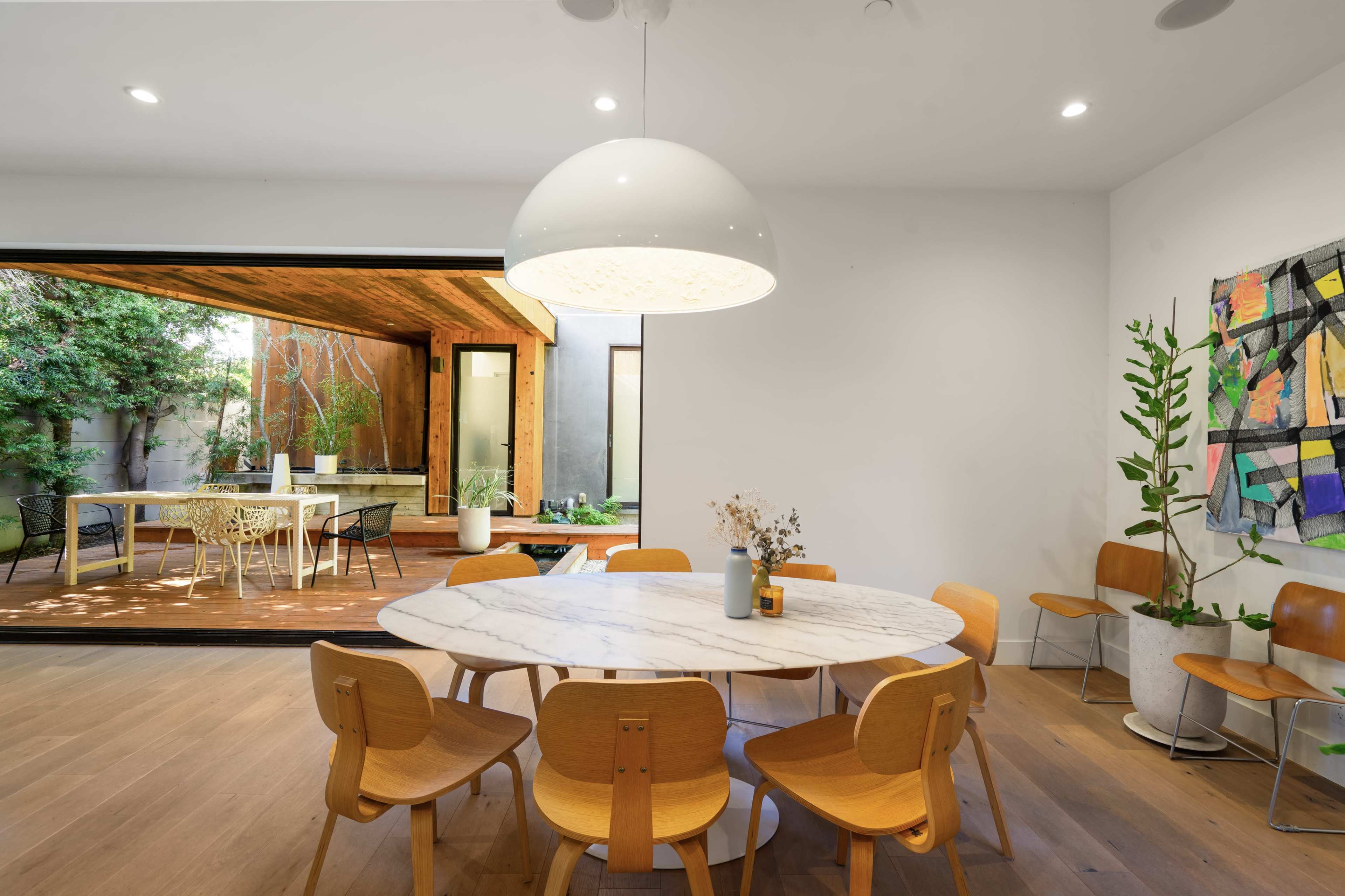 A round marble dining table surrounded by wooden chairs sits in a well-lit room that opens to an outdoor patio area with greenery.
