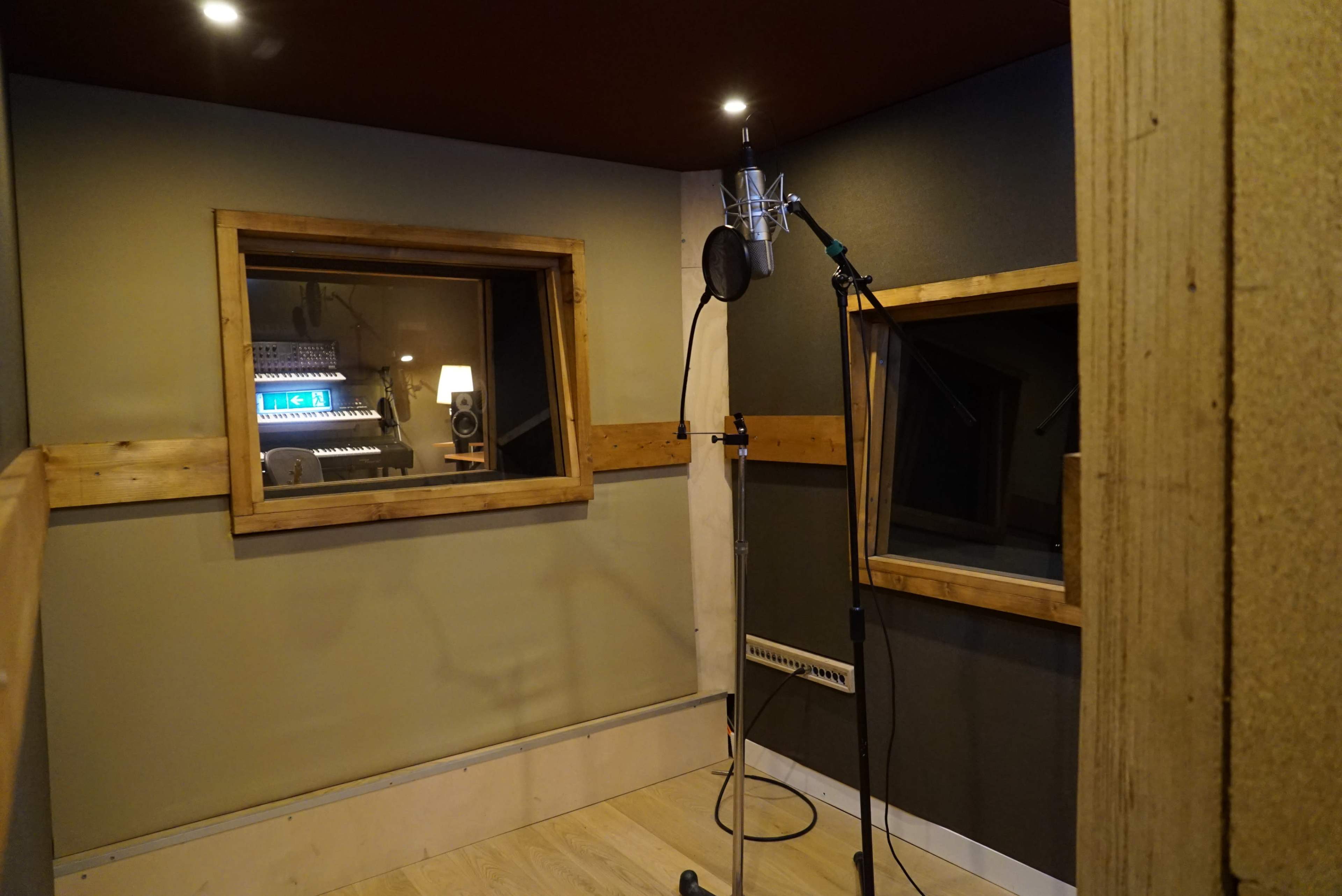 A recording studio with a microphone on a stand, two windows for sound isolation, and a control panel visible through one of the windows.