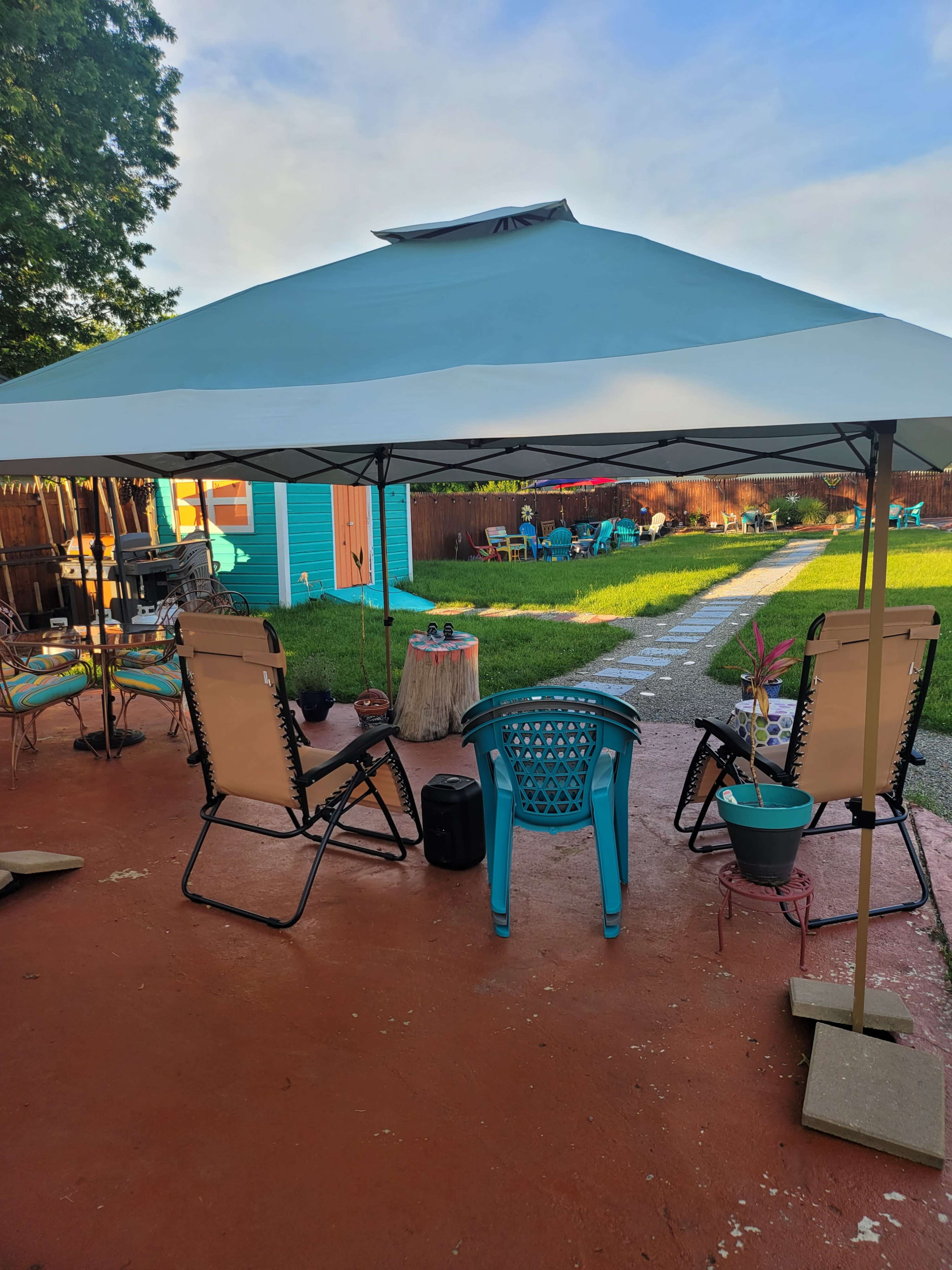 A shaded seating area with two lounge chairs, a blue side table, and a potted plant overlooks a well-kept backyard with a pathway and colorful outdoor furniture.