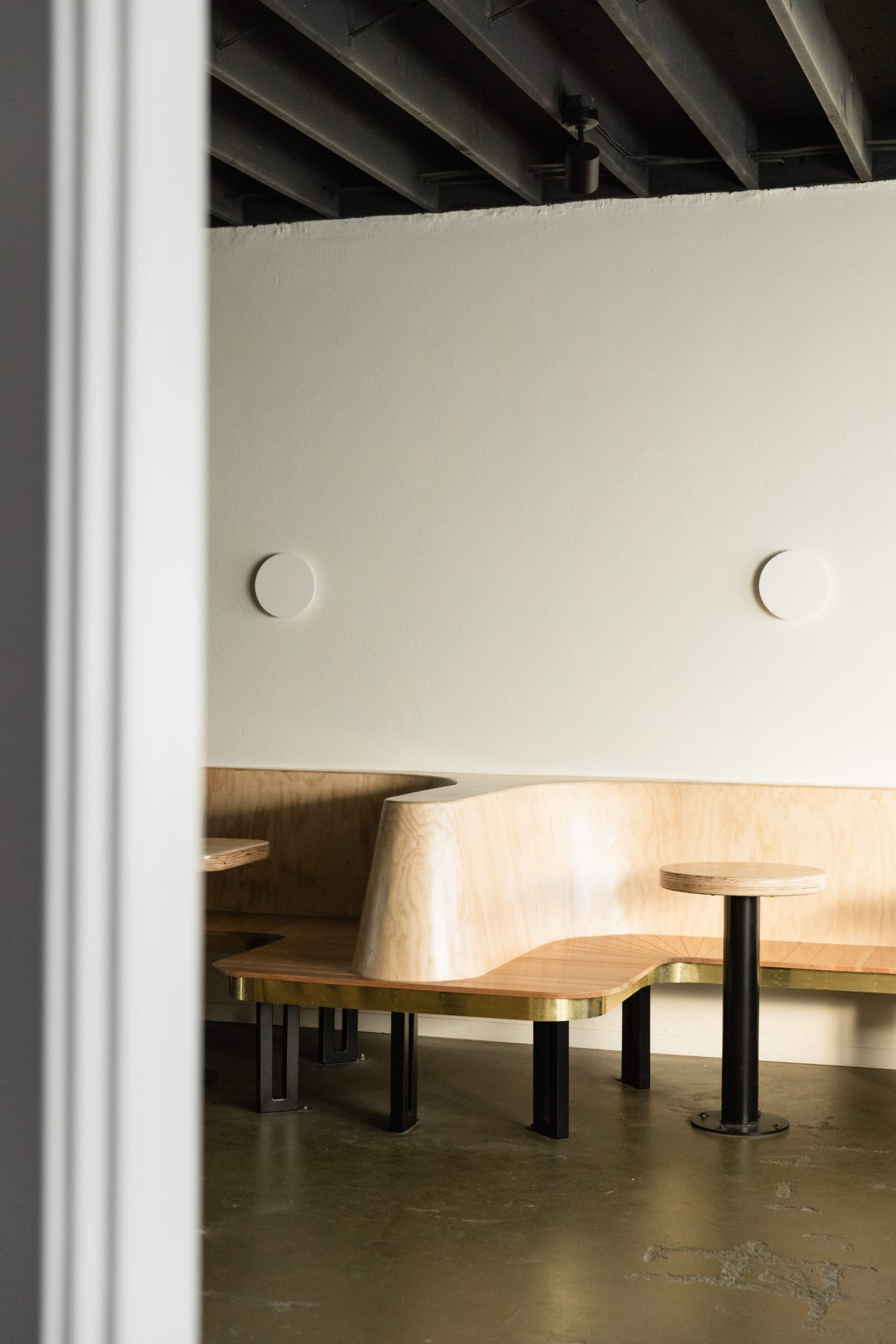 The image shows a minimalist interior with curved wooden seating and black tables against a plain wall.