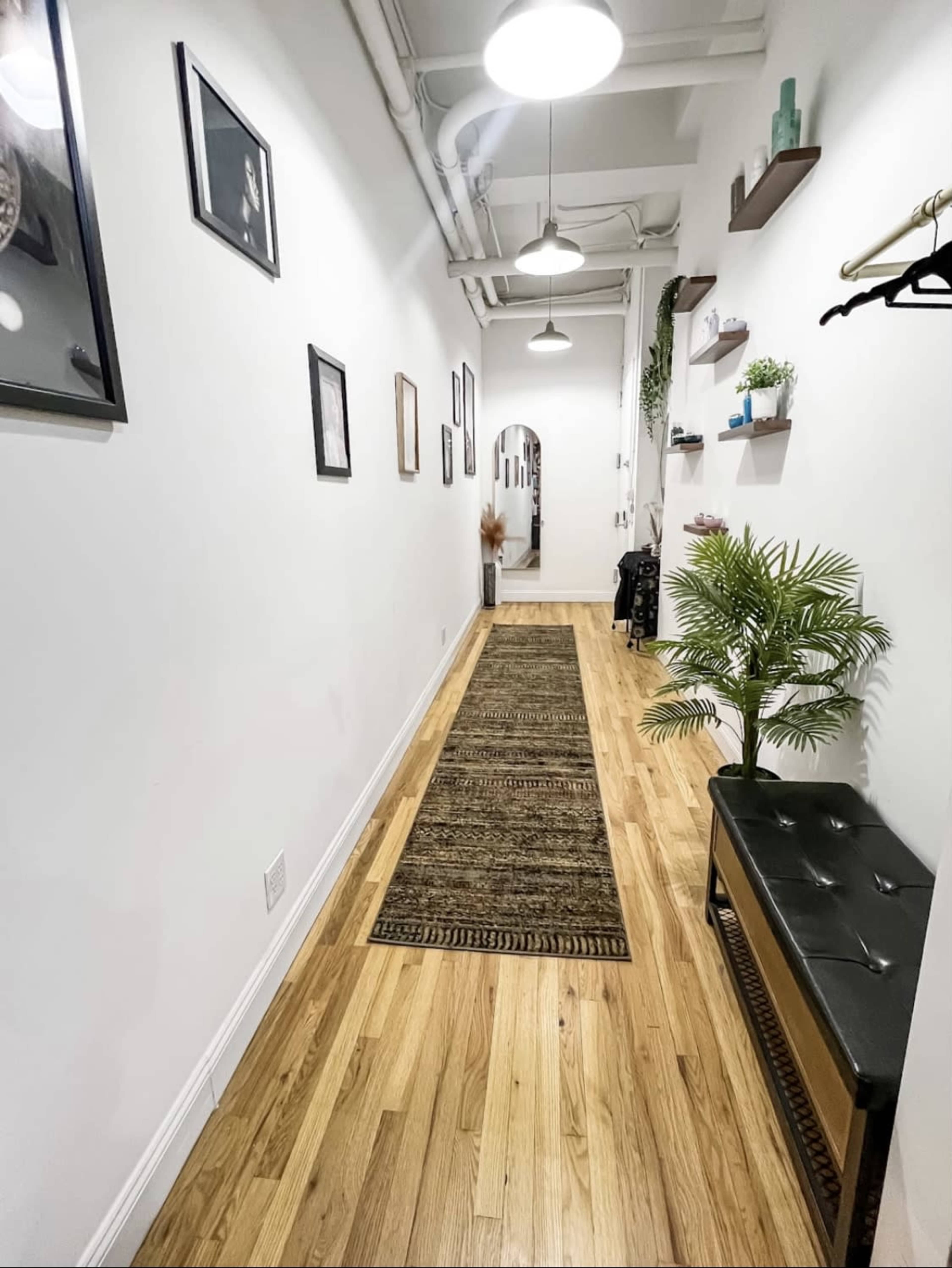 A long, narrow hallway with wooden flooring, a runner rug, framed pictures on the walls, and a decorative plant beside a small bench.