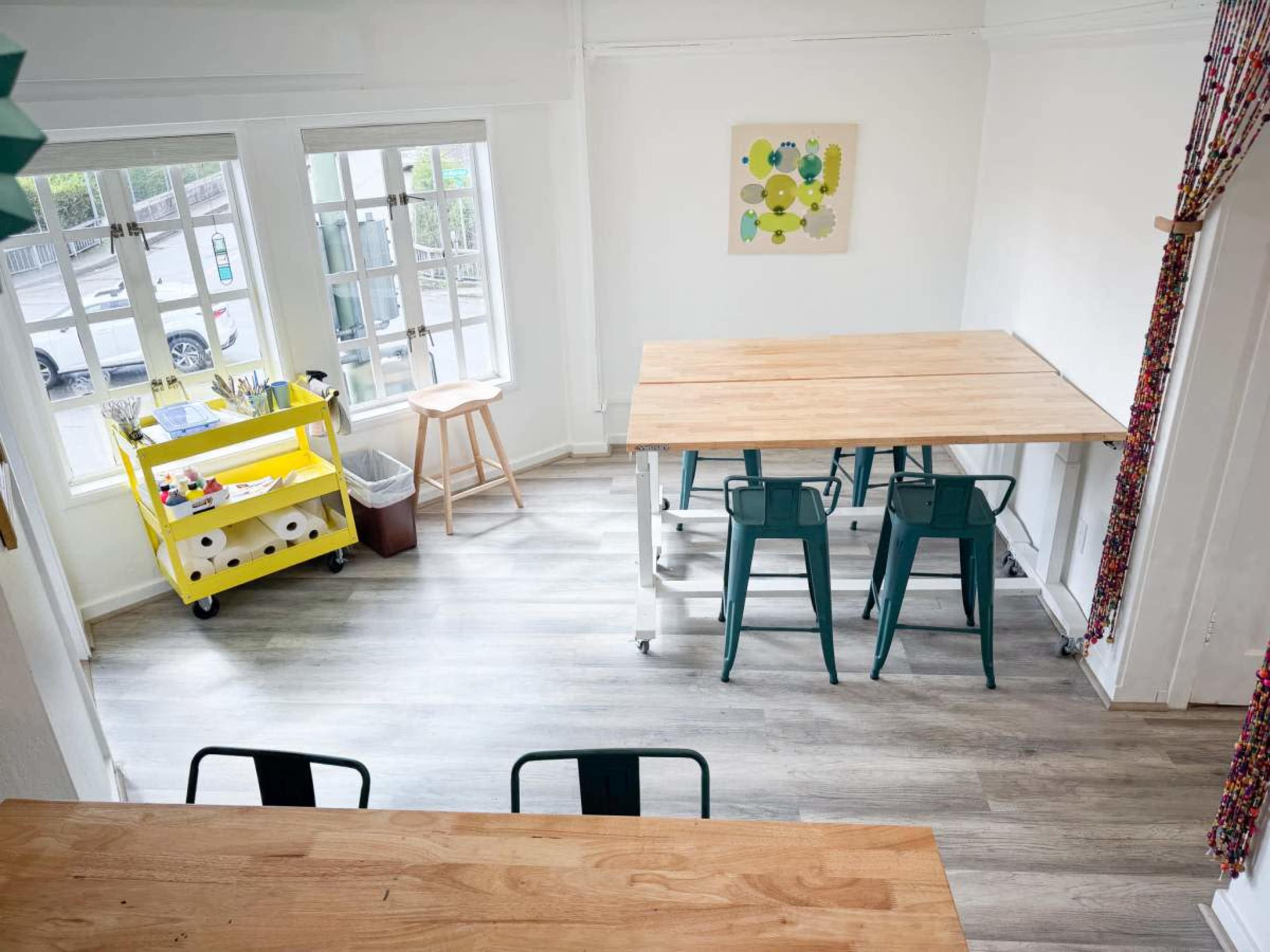 A bright, spacious room with a large wooden table, green metal chairs, and a yellow cart filled with supplies.