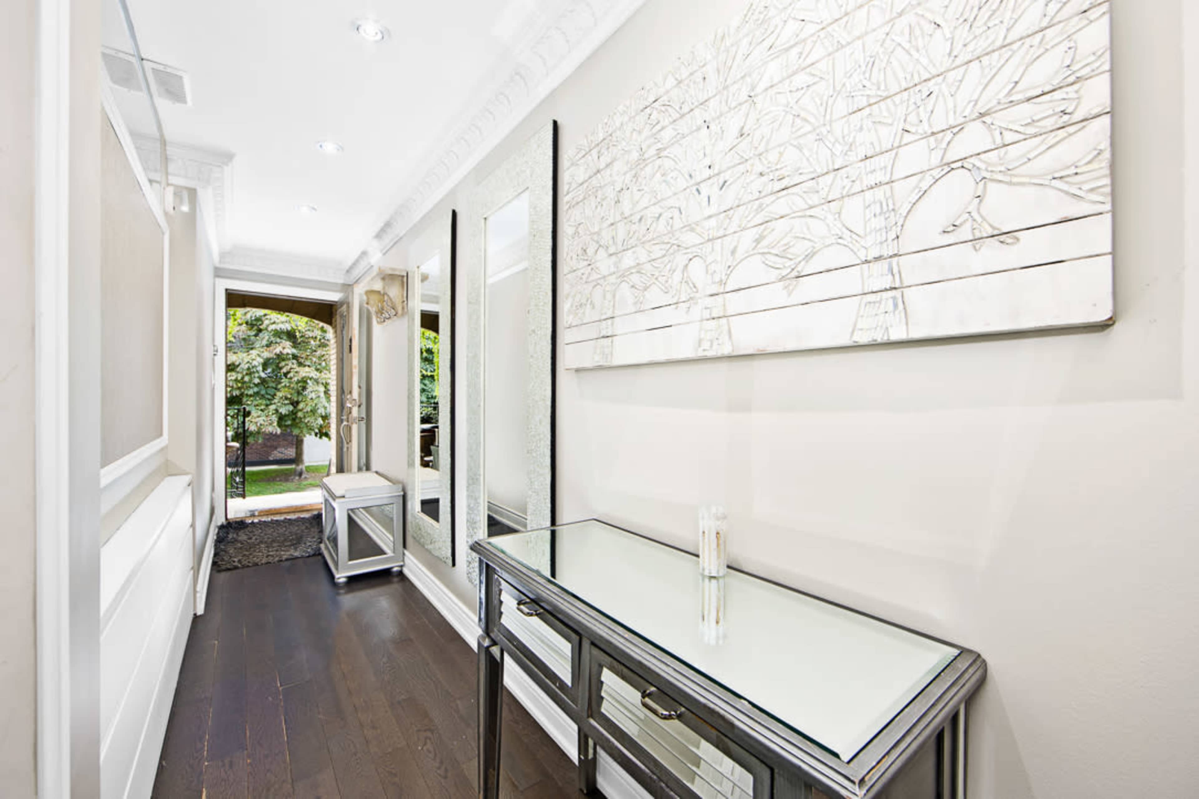 The image shows a sleek hallway with a console table, a large mirror, and a decorative wall art featuring tree designs, leading to an entrance that opens to an outdoor area.