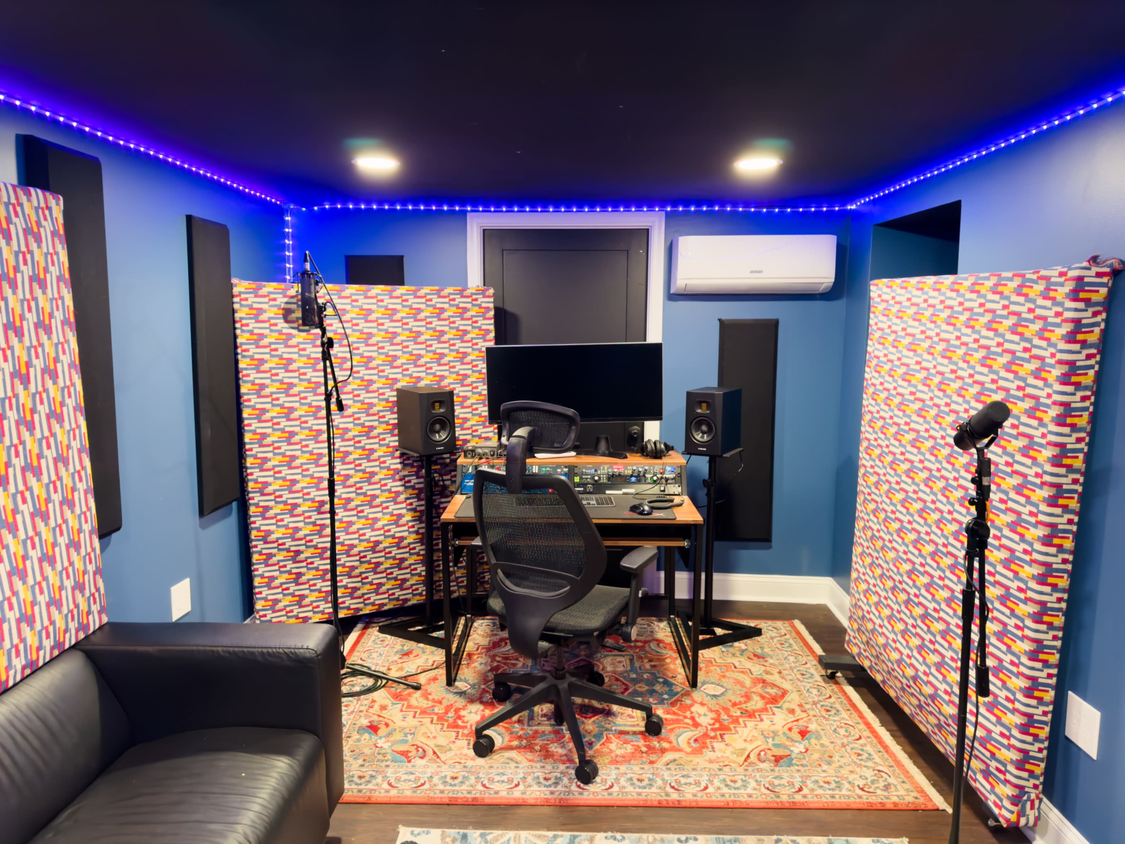 A small recording studio equipped with a desk, audio monitors, a microphone setup, and sound-absorbing panels.