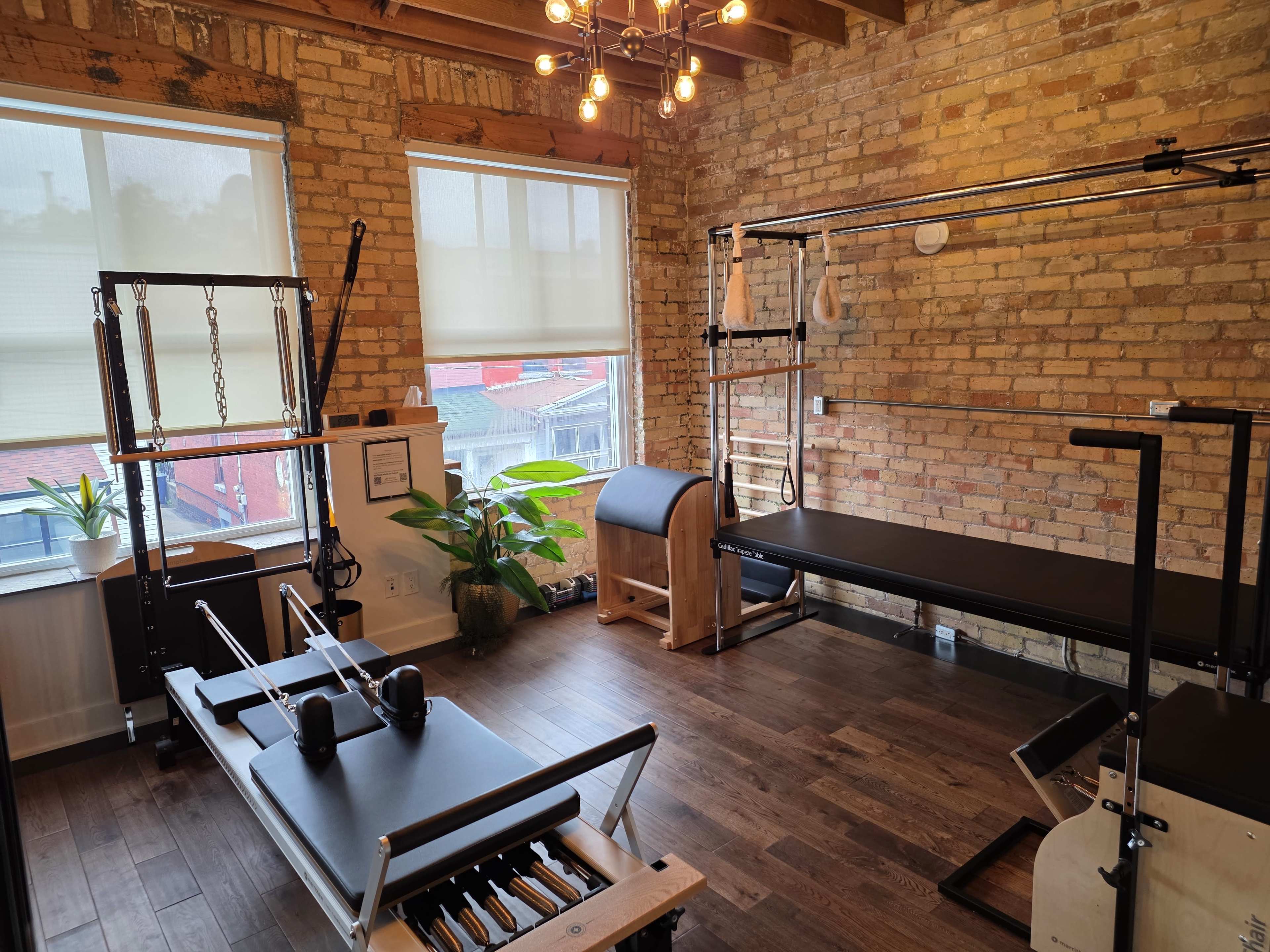 The image shows a well-lit Pilates studio with wooden floors, brick walls, and various exercise equipment, including a reformer, a barrel, and a wall-mounted apparatus.