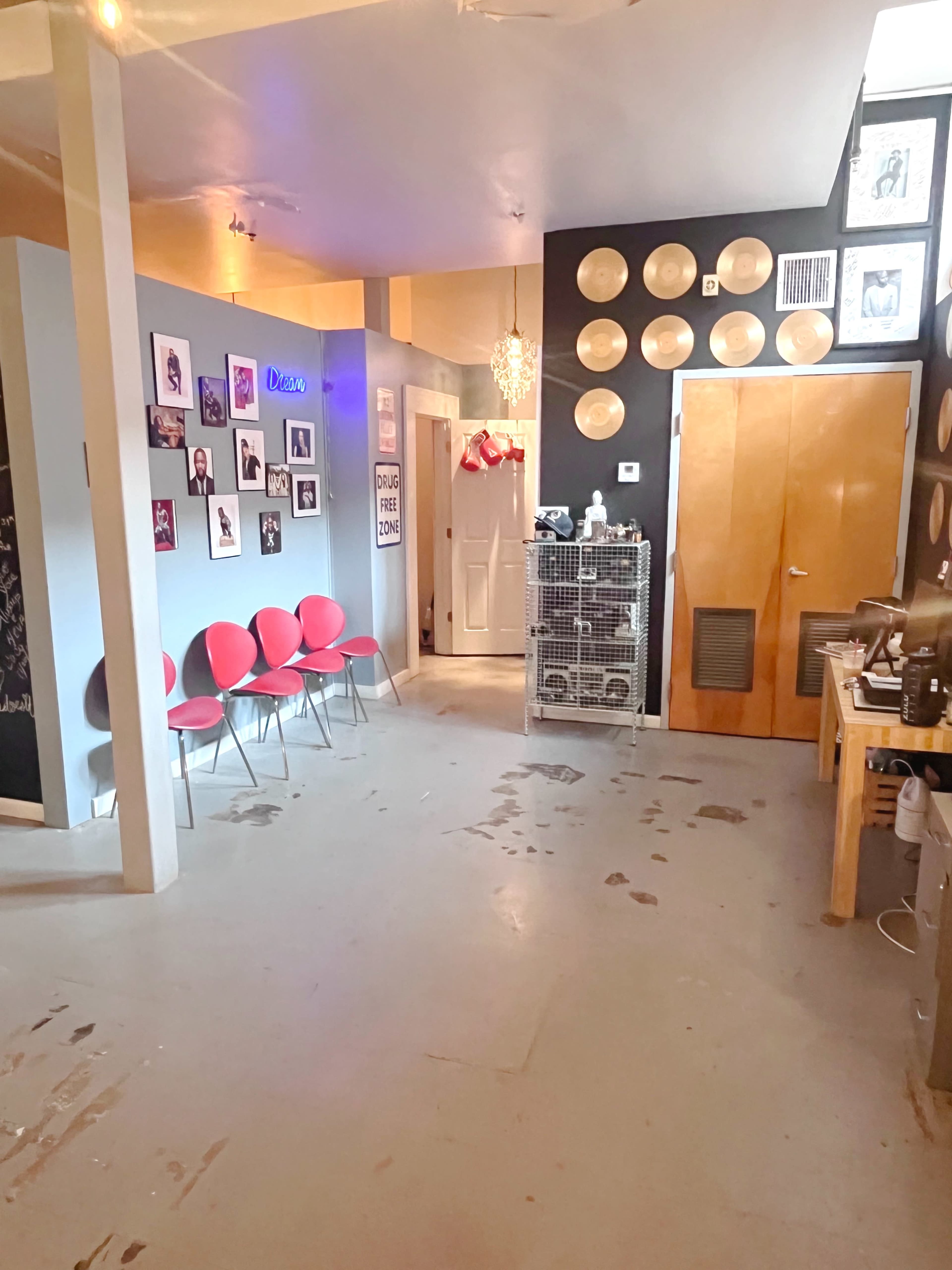 The image shows a room with a polished floor, featuring red chairs along one wall, framed photos on another, and a door leading to a hallway.