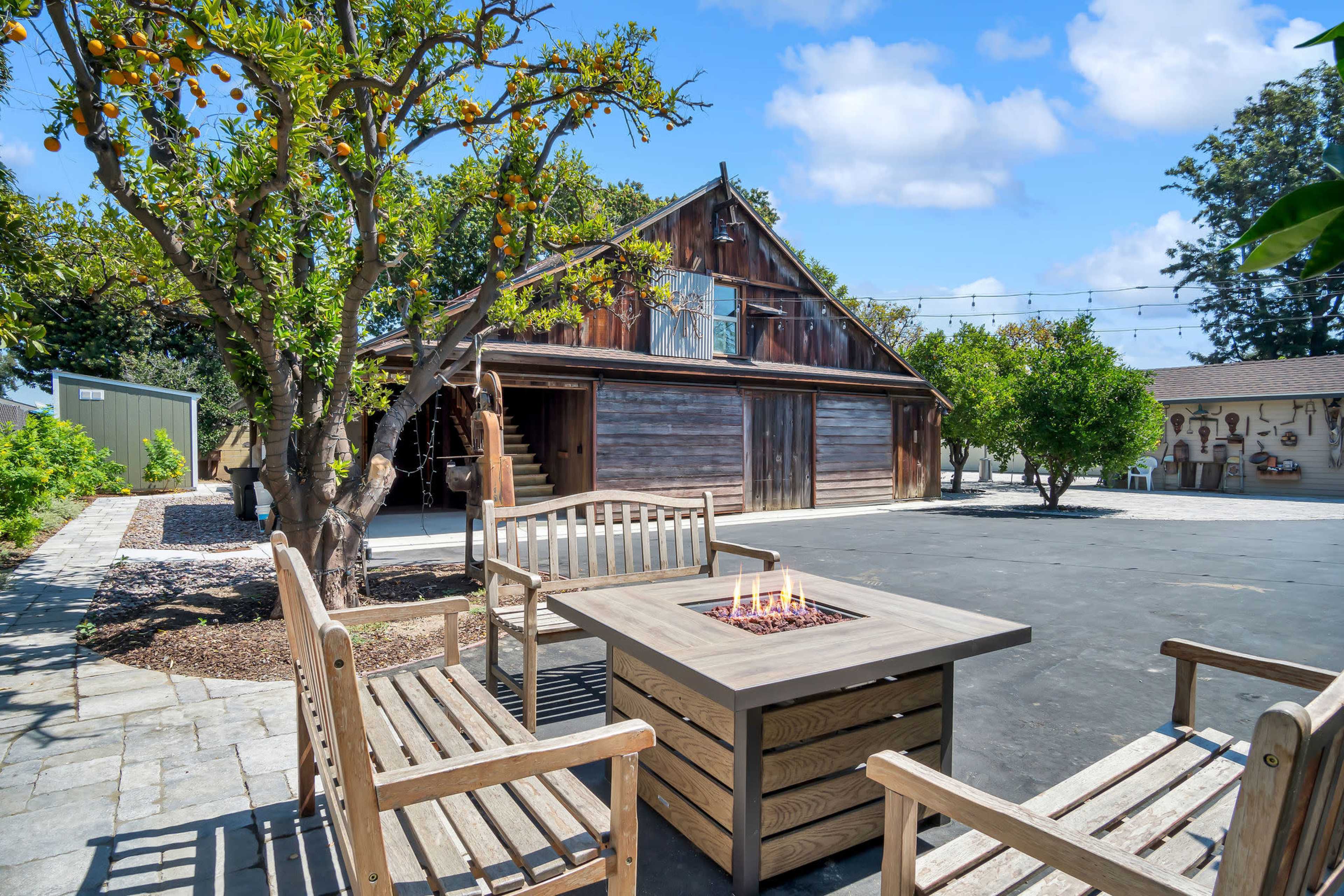 A rustic wooden building serves as a backdrop for a patio area featuring a fire pit and benches, surrounded by trees and landscaped greenery.