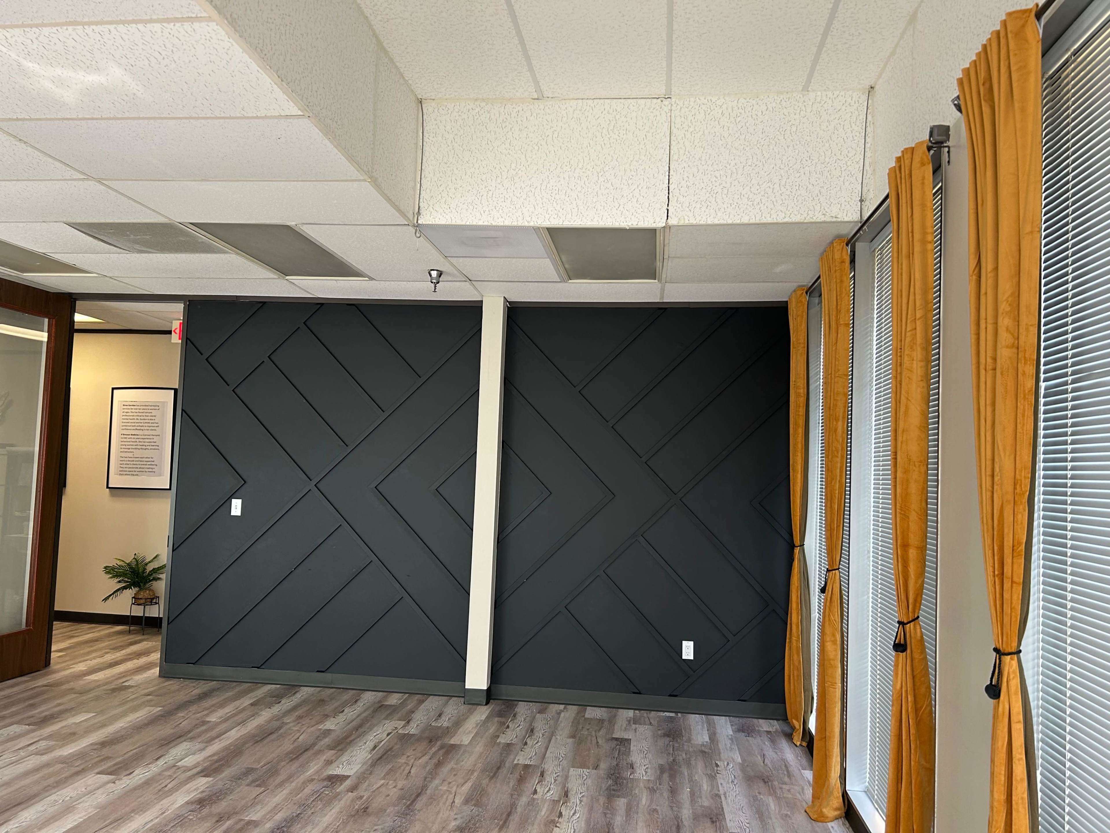 The image shows an interior workspace with a dark geometric patterned wall and large windows covered by orange curtains.