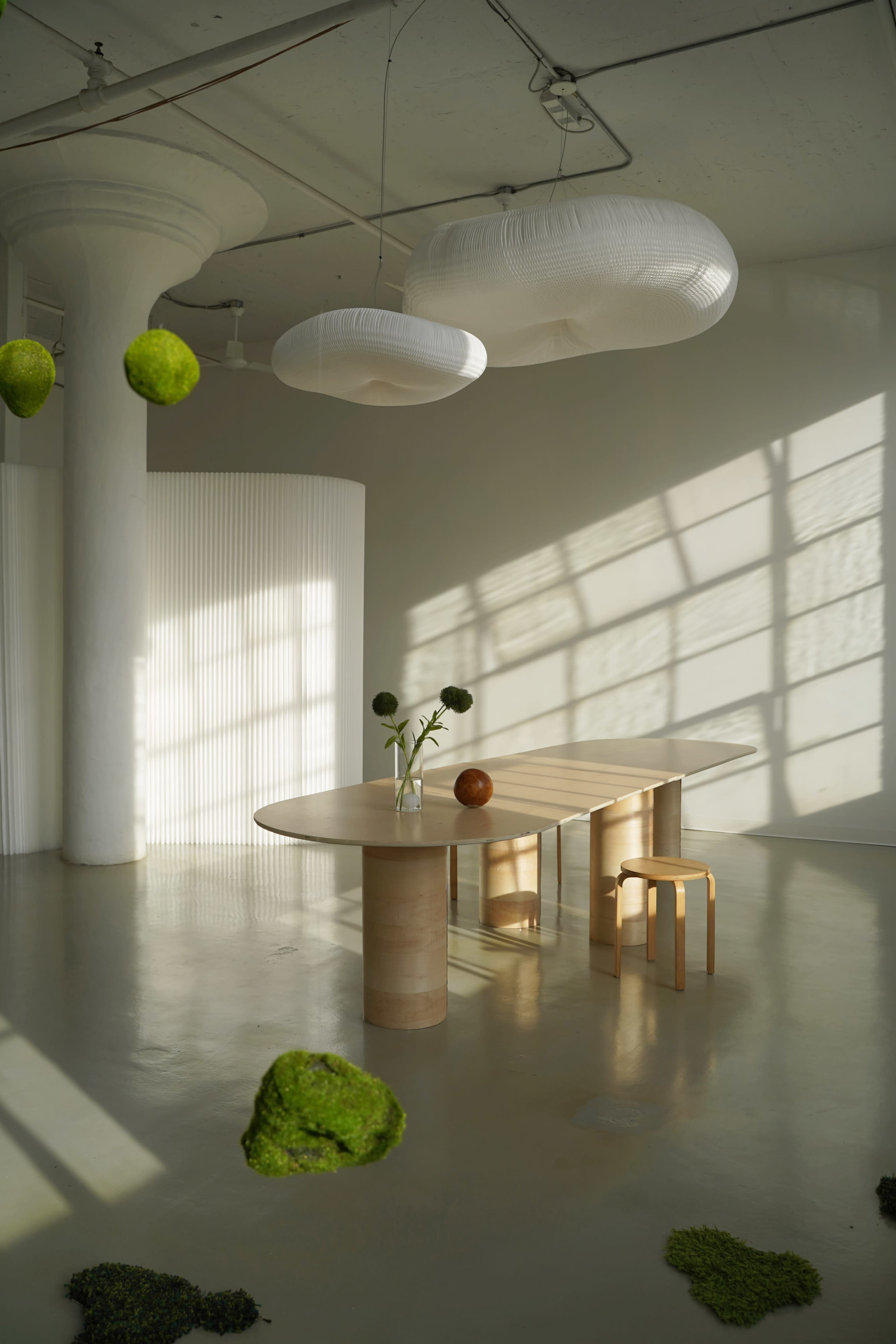 A minimalist dining area features a round wooden table, a clear vase with flowers, and floating light fixtures casting shadows on the smooth floor.