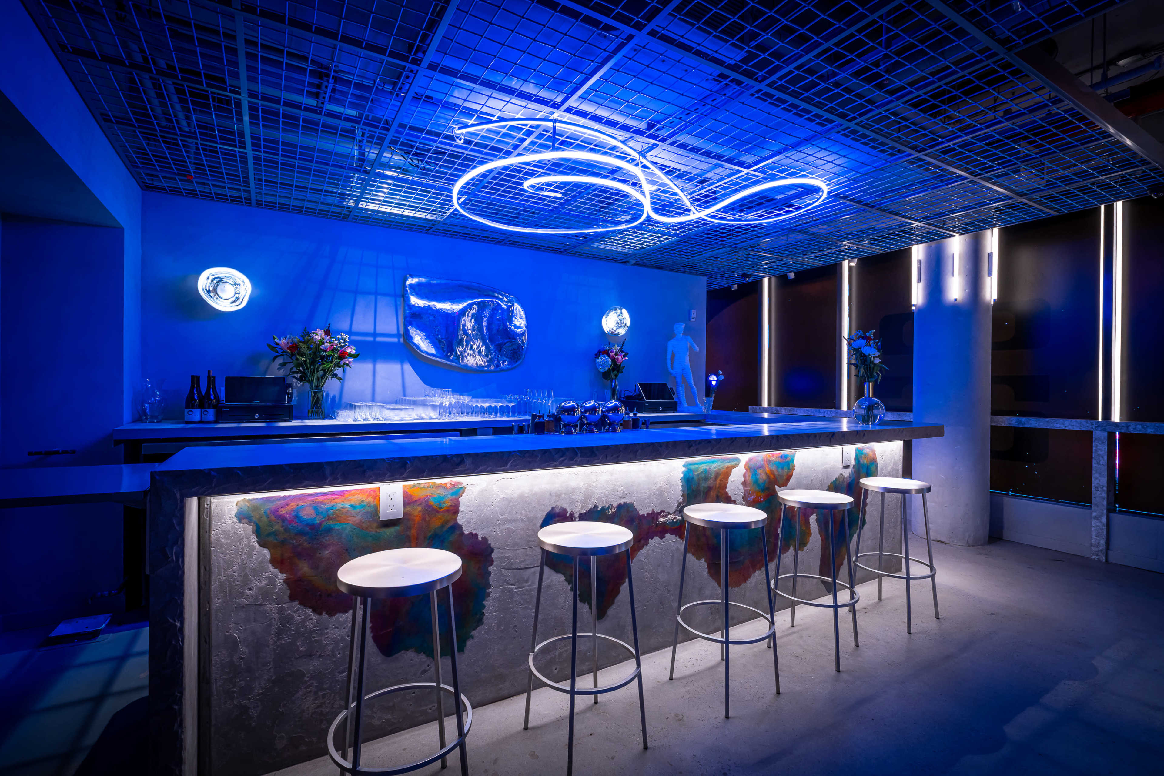 A modern bar with a blue neon light overhead, sleek countertops, and several stools arranged in front.