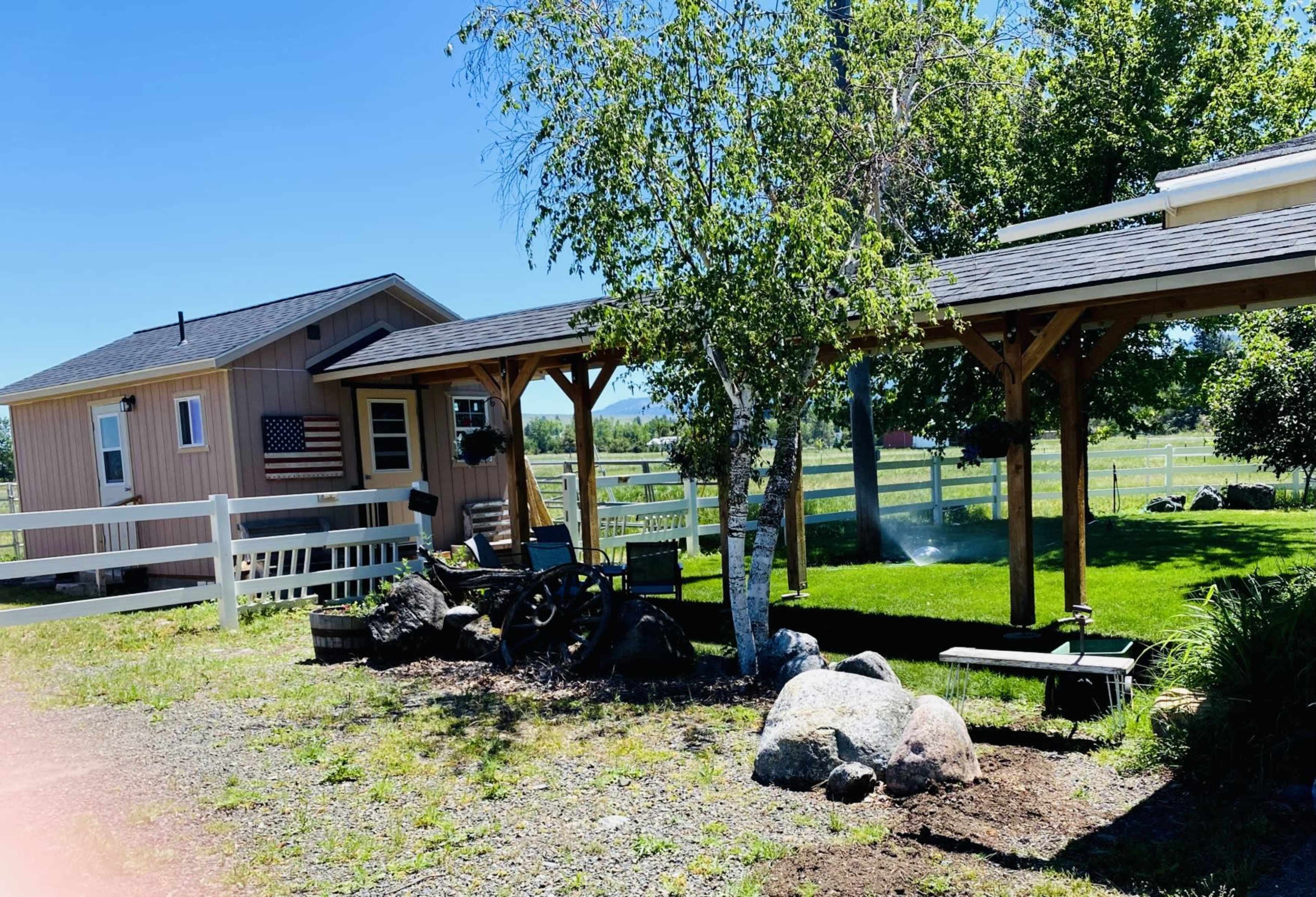 A small cabin with an American flag and a shaded seating area is set among rocks and greenery in a rural landscape.