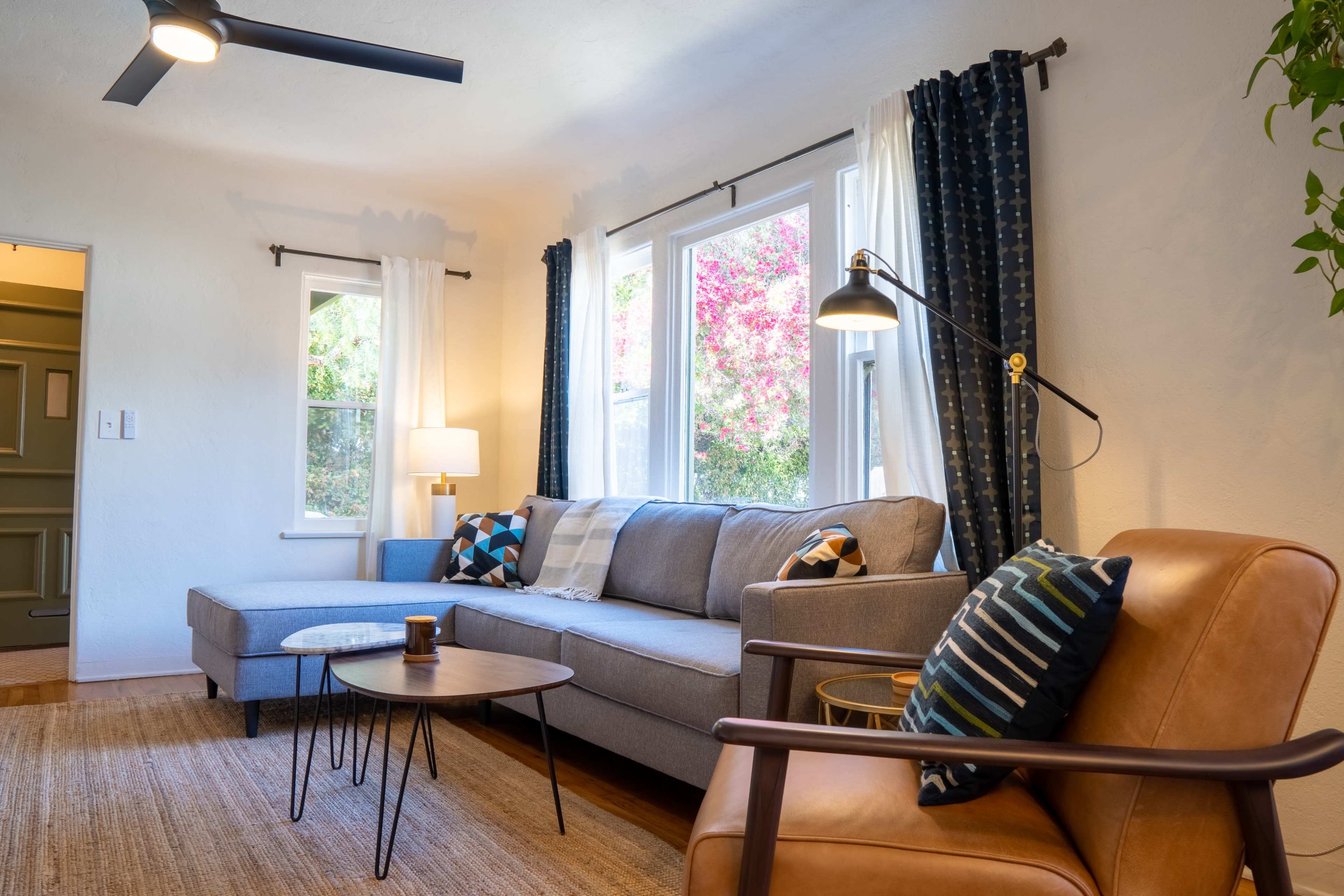 A living room features a grey sectional sofa, a wooden coffee table, two windows with sheer curtains, and a brown armchair beside a decorative lamp.