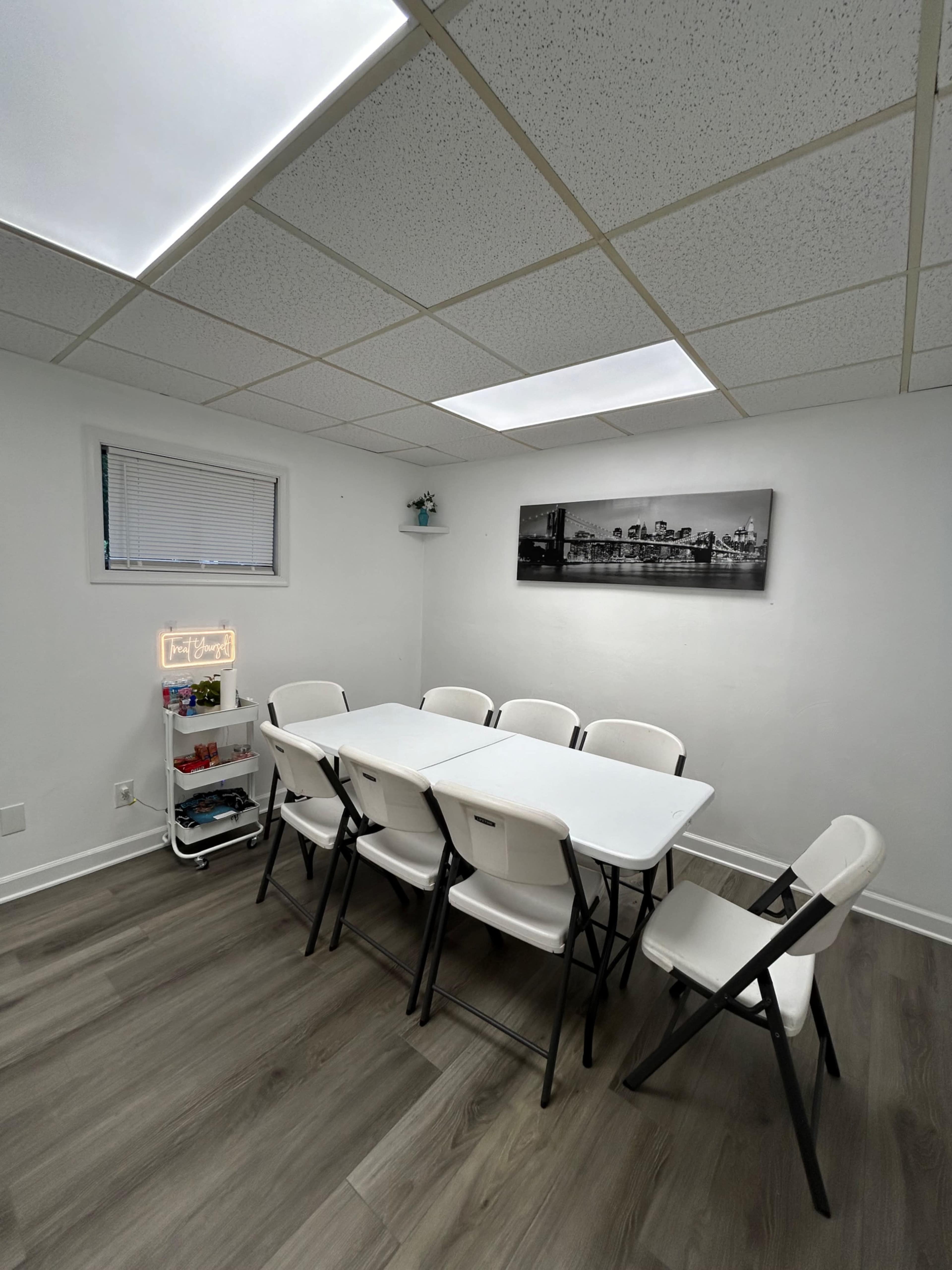 A simple meeting room features a long white table surrounded by six folding chairs, with a small shelf holding books in one corner and a black-and-white cityscape photograph on the wall.