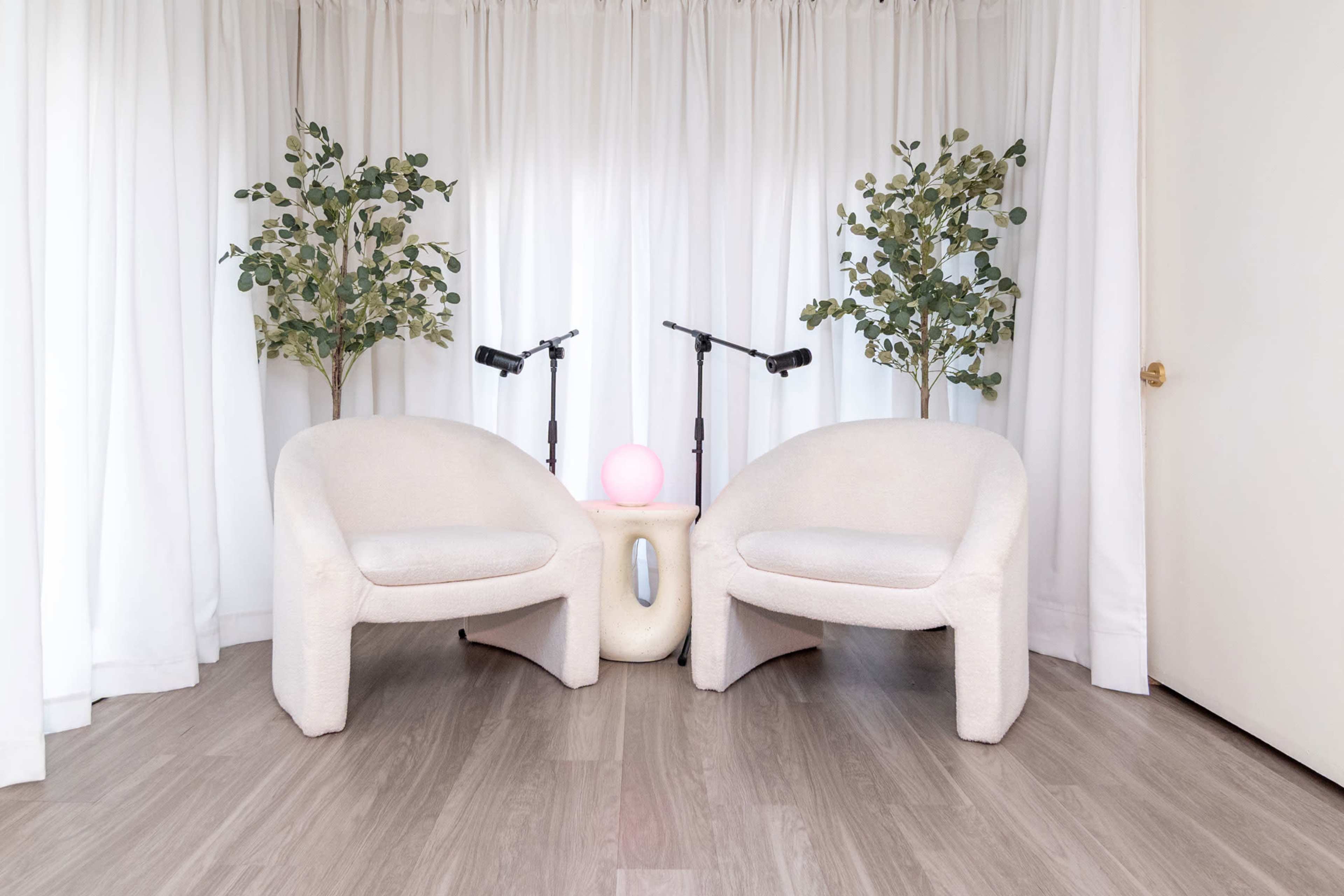 Two beige chairs are positioned facing each other beside a round pink light on a sculptural table, with green plants flanking them and sheer white curtains in the background.