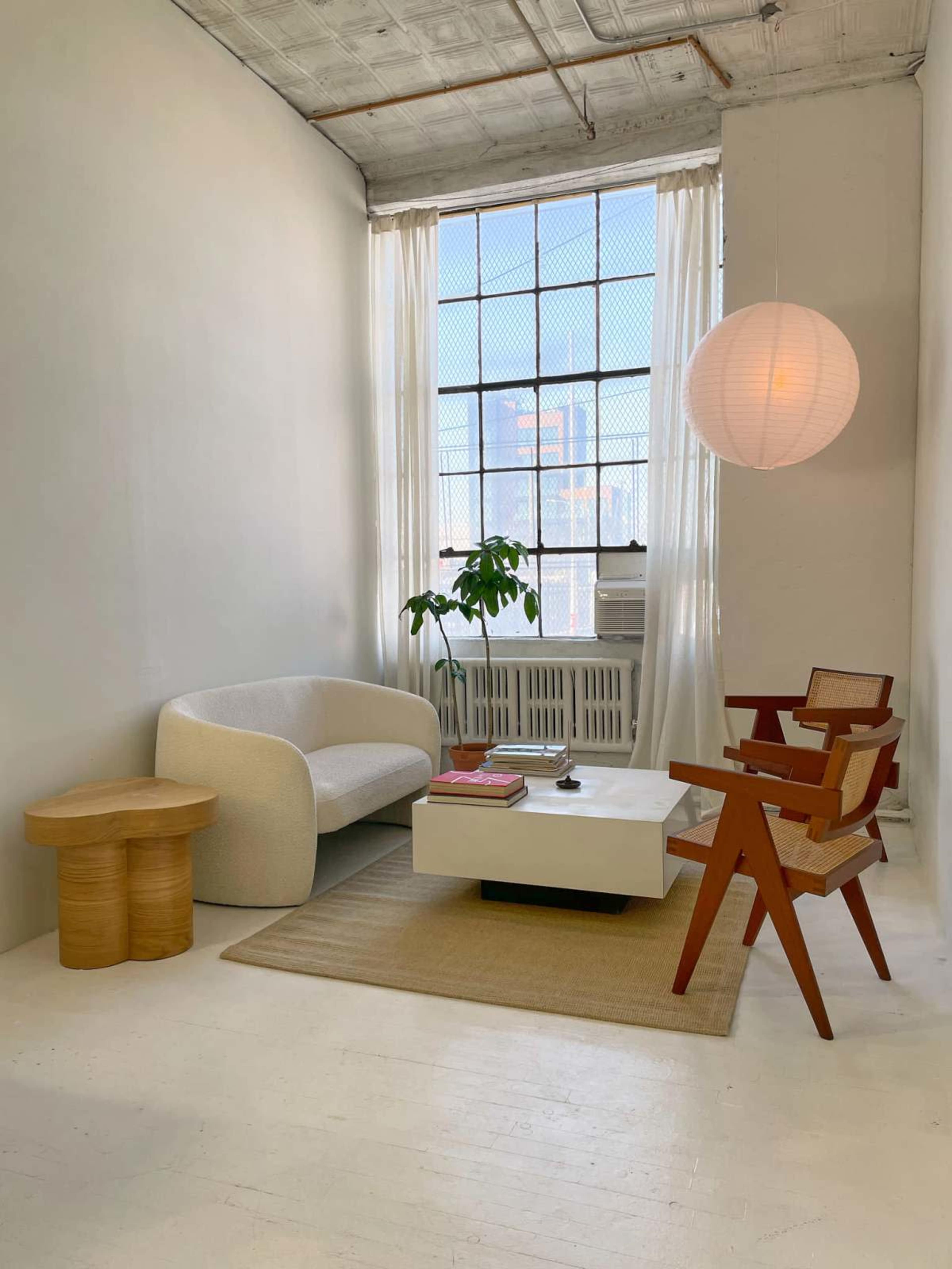 The image shows a minimalist living room with a light-colored sofa, a wooden side table, a white coffee table, and a hanging paper lantern, all arranged near a large window with sheer curtains.