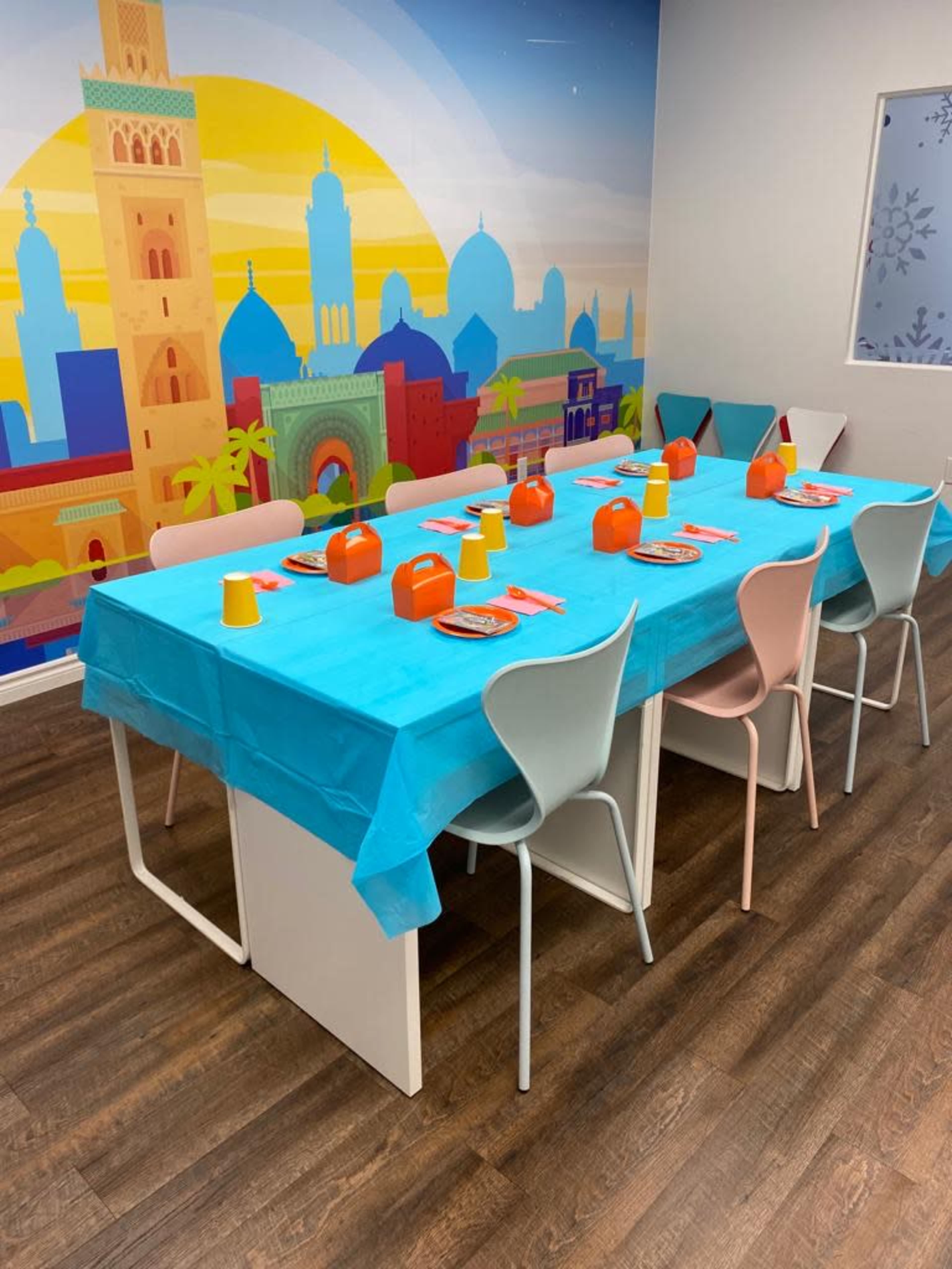 A large table is set with a blue tablecloth, colorful cups, and plates, surrounded by chairs in a room adorned with a vibrant mural.