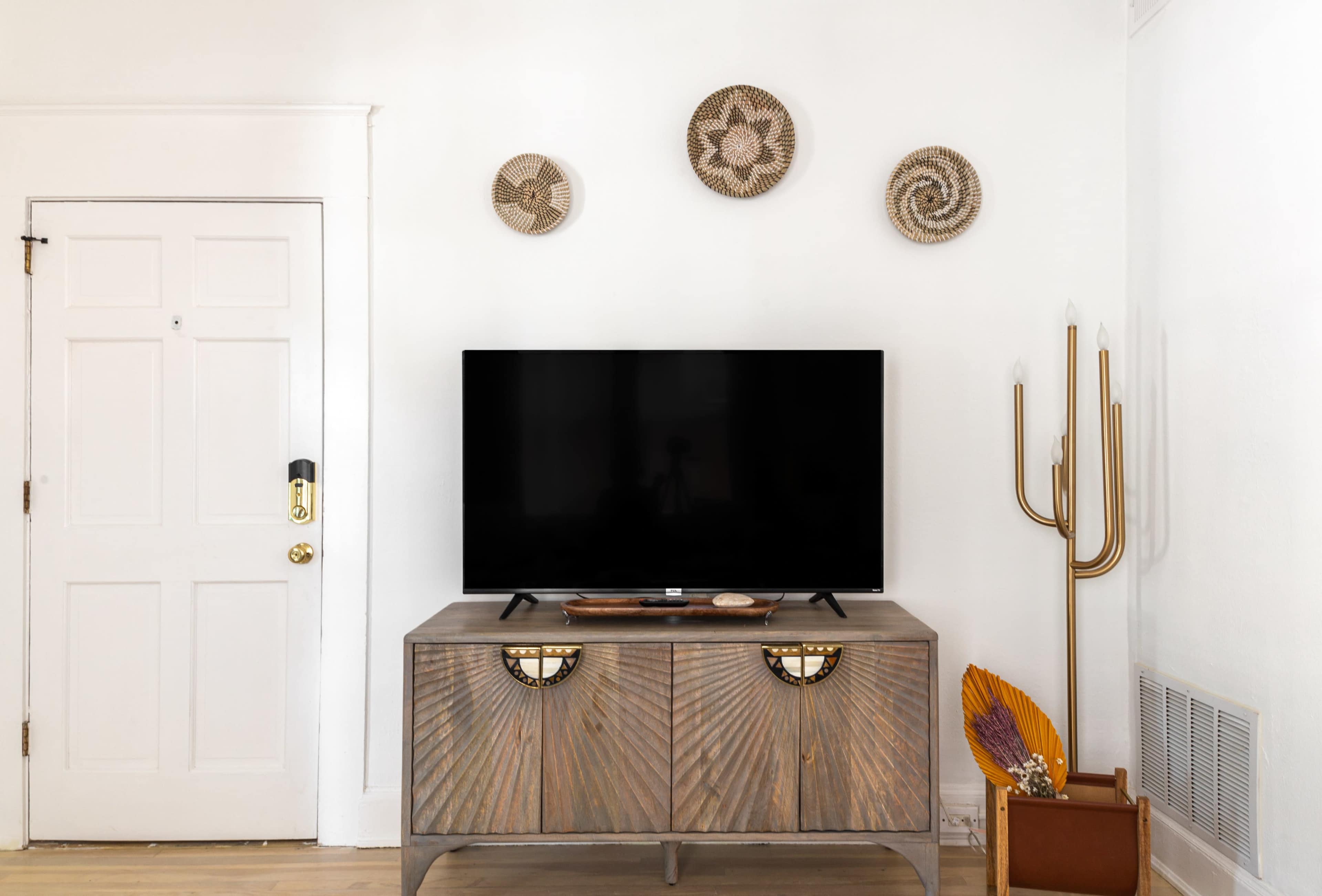 A wall-mounted television is placed above a wooden console table with decorative elements, and three woven wall hangings are arranged above it.