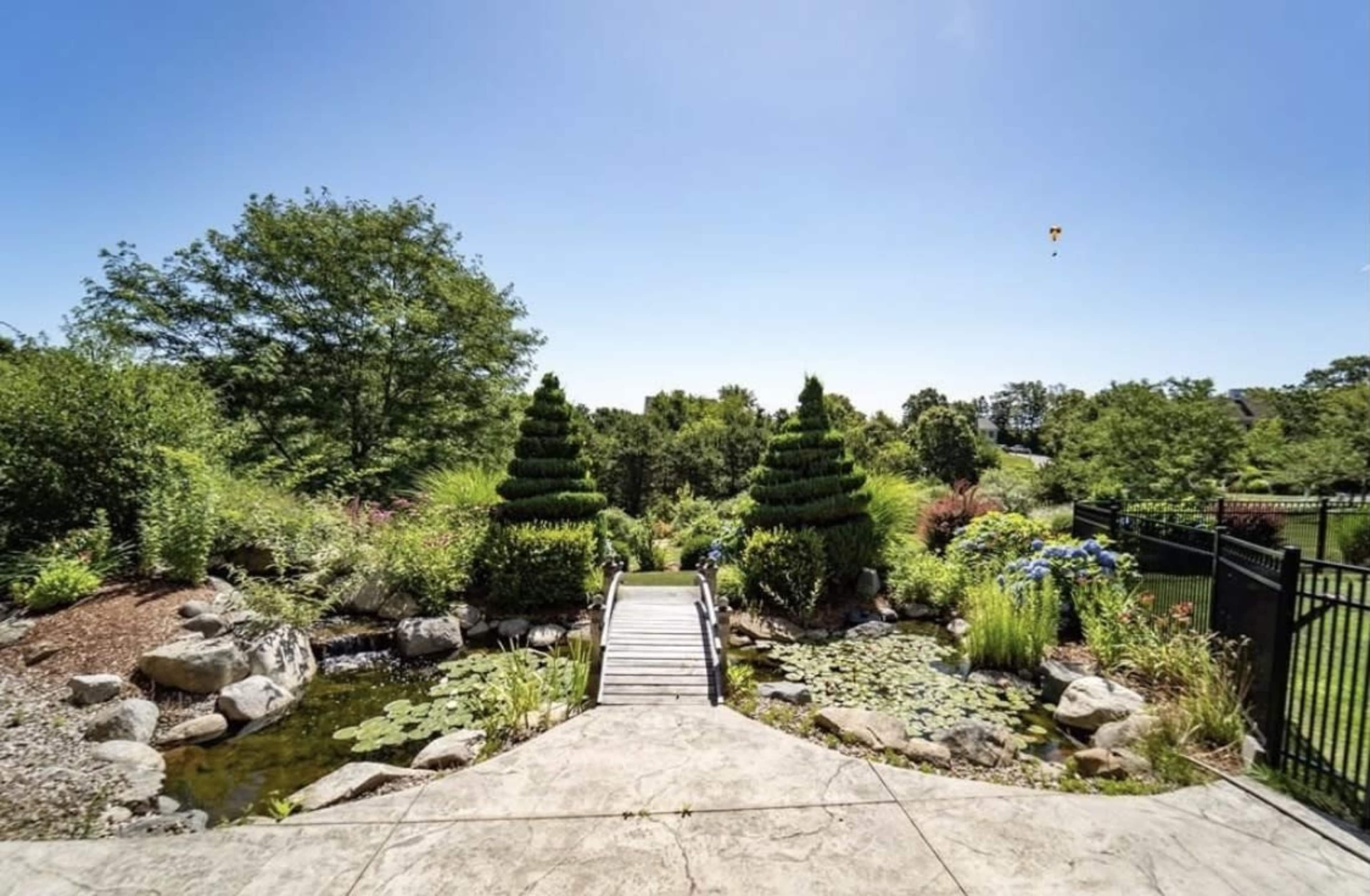 A wooden bridge leads over a pond surrounded by manicured greenery and topiary trees against a clear blue sky.